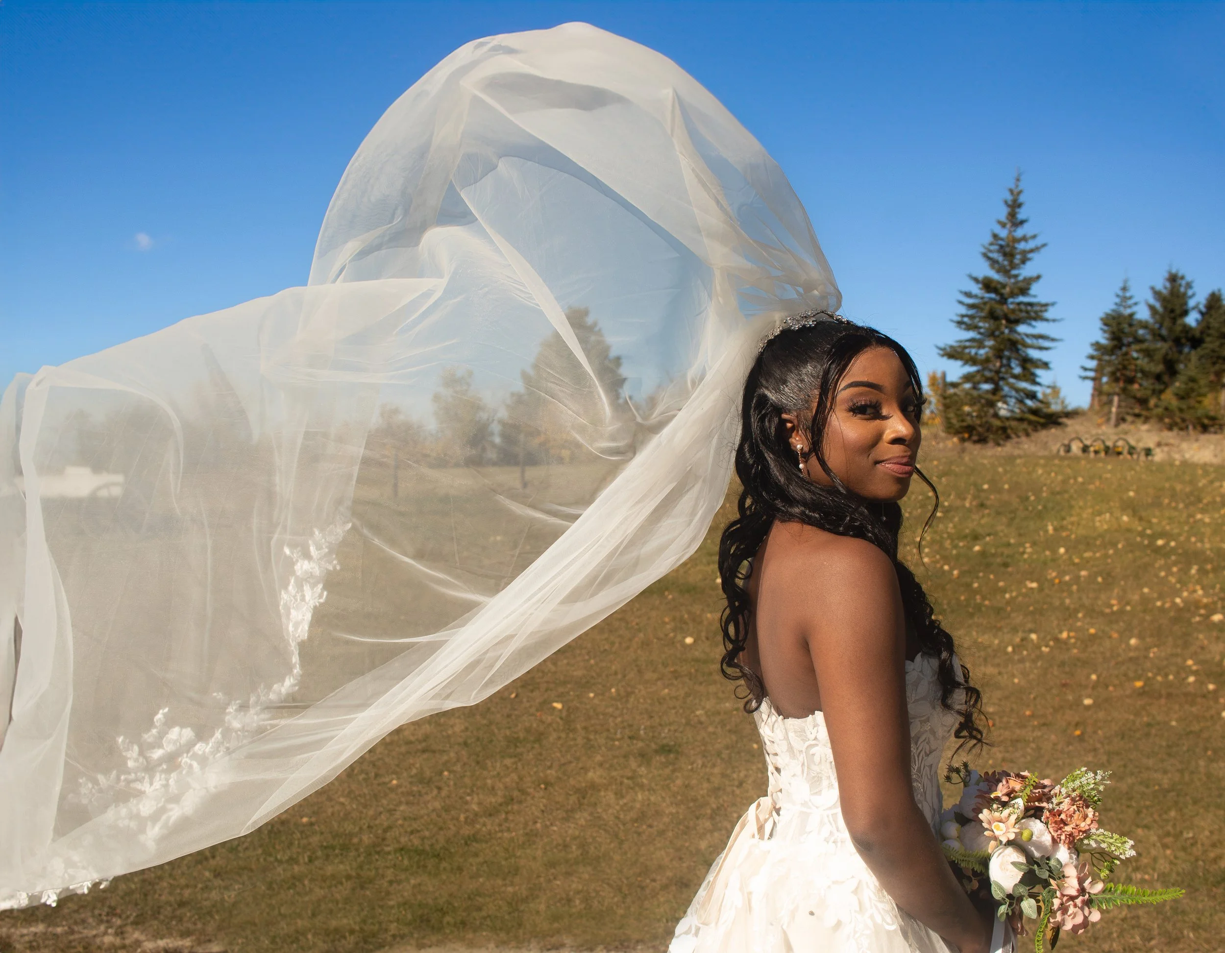 A woman wearing a wedding dress and veil outdoors, holding a bouquet of flowers, with trees and a clear blue sky in the background.