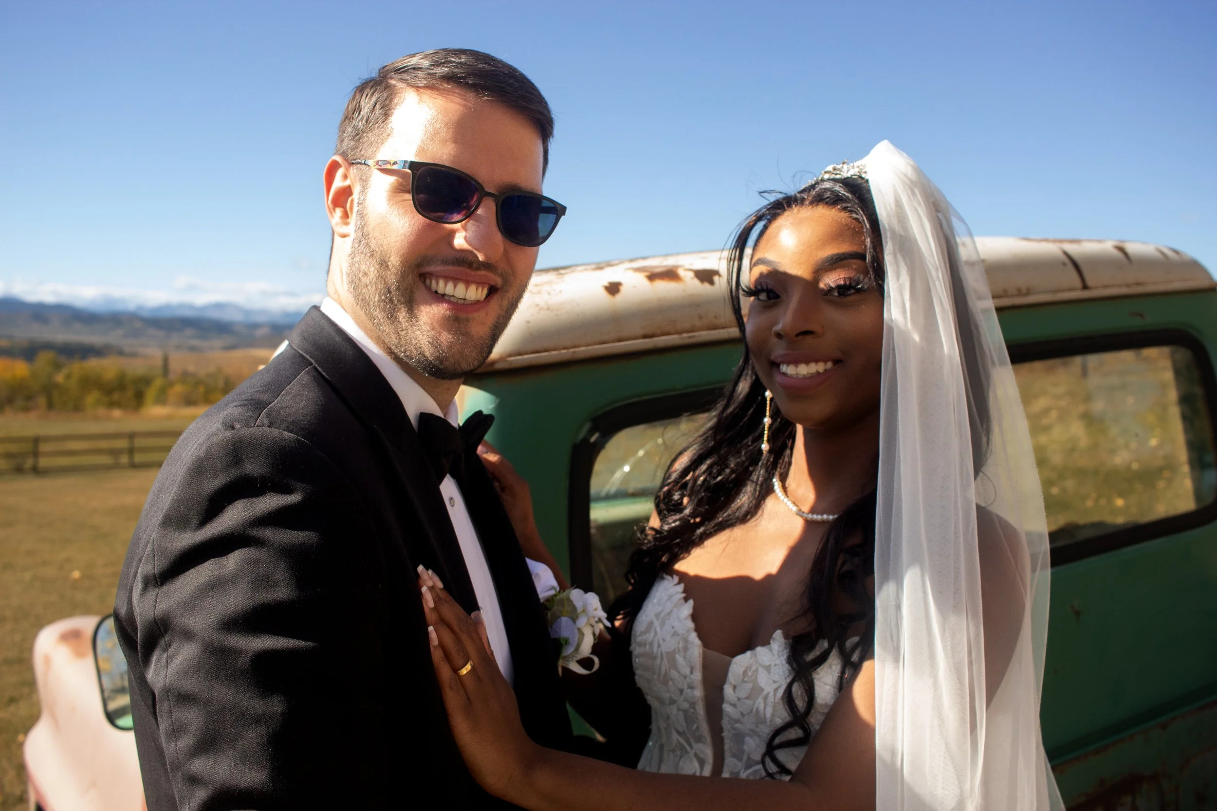 A wedding couple smiling, standing outdoors with a vintage green vehicle and a rural landscape in the background.