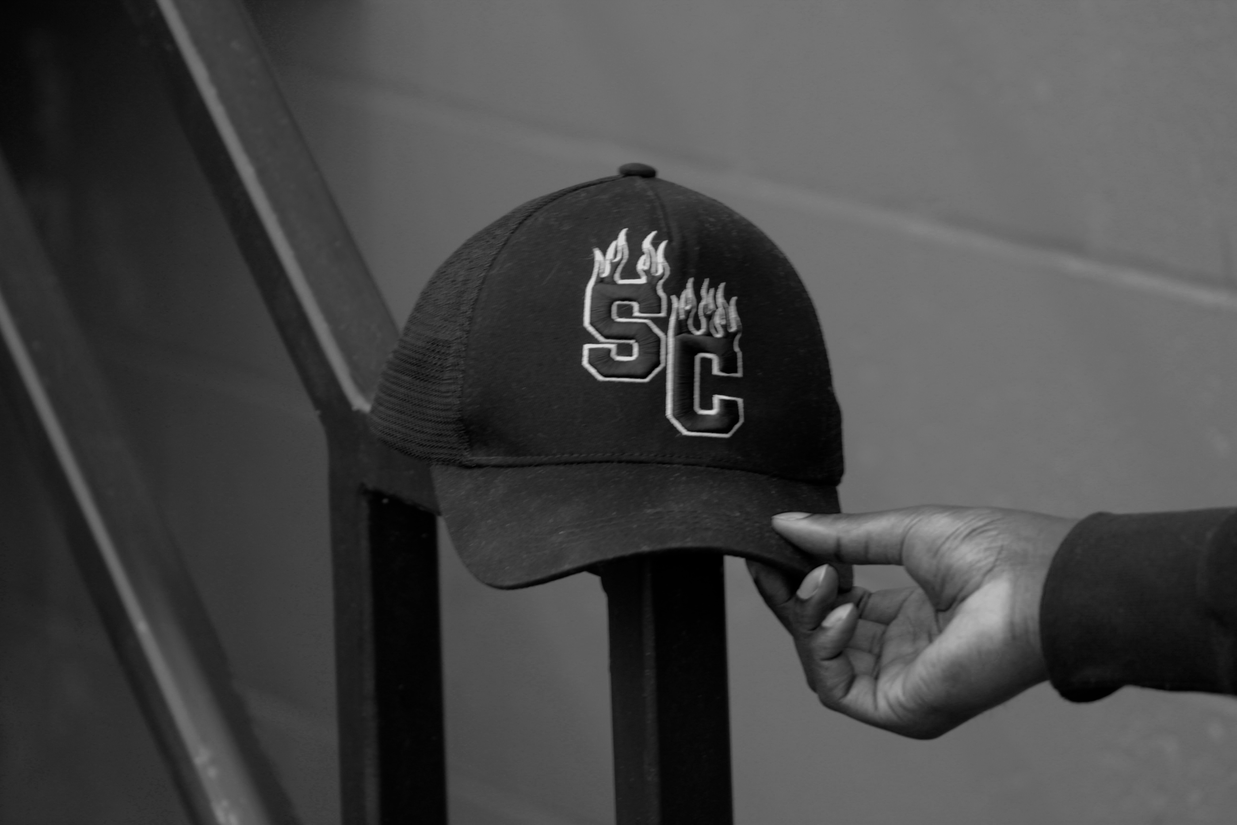 Black and white photo of a person’s hand holding a black baseball cap with the letter 'S' and additional design on the front, near a mirror or reflective surface, with part of their hand and a dark object visible in the frame.