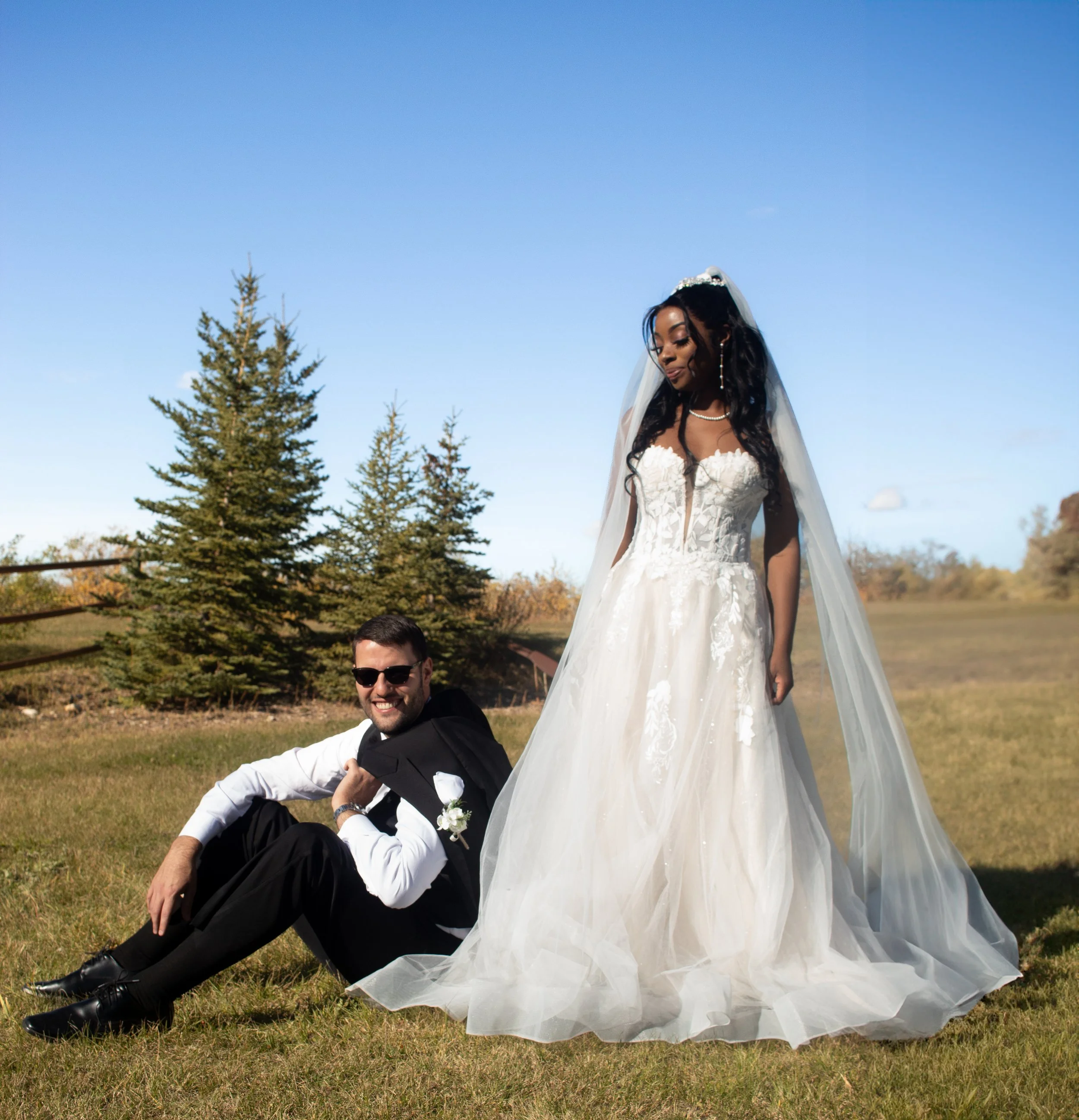A bride in a white wedding dress and veil looking down, standing outdoors on grass with trees in the background, with a man in a tuxedo sitting on the ground smiling.