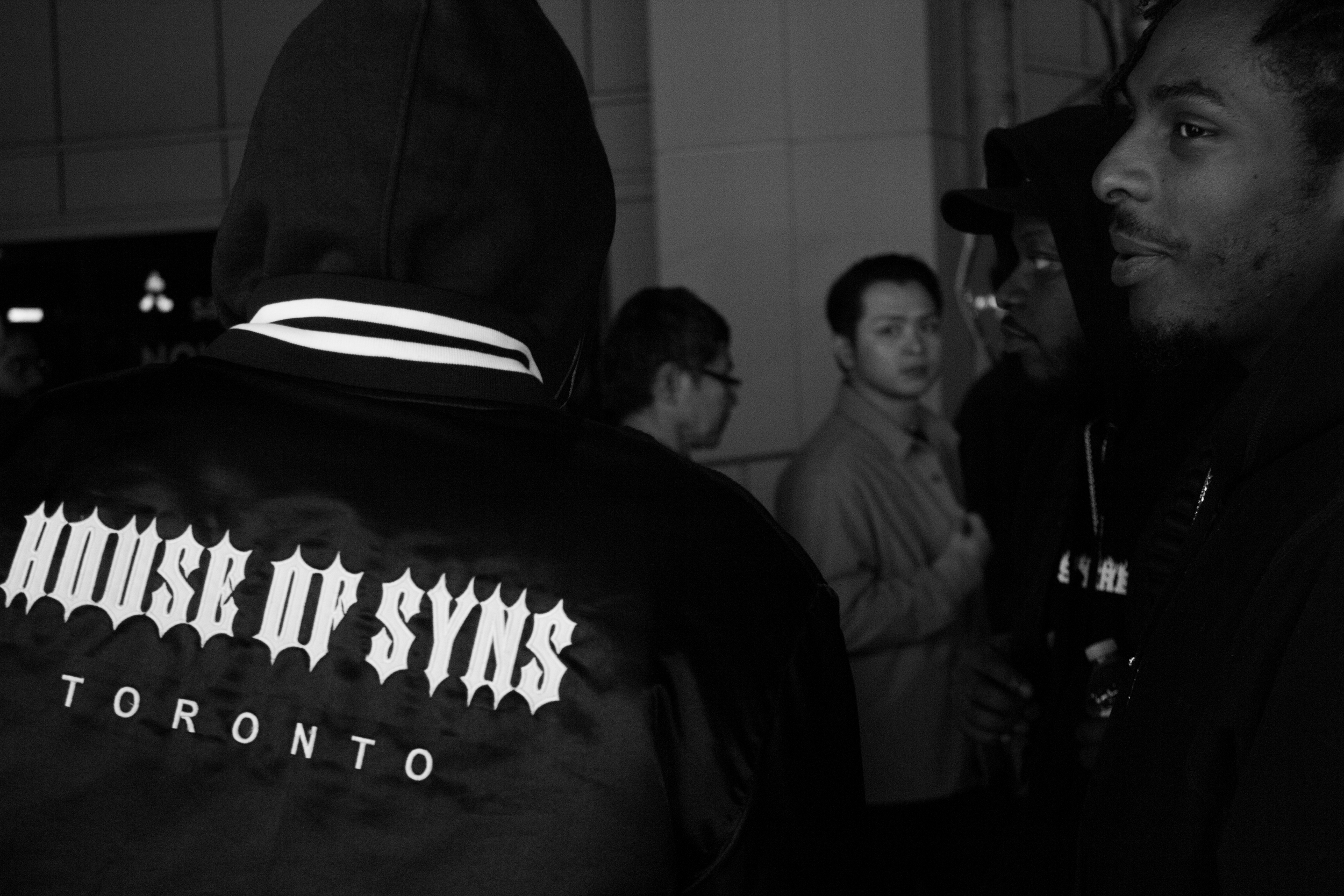 People gathered together in a public indoor space, some wearing hoodies and jackets, with focus on a man with dark skin and short hair facing others, background features plain walls and in-focus individuals, one with glasses and another with a cap, e