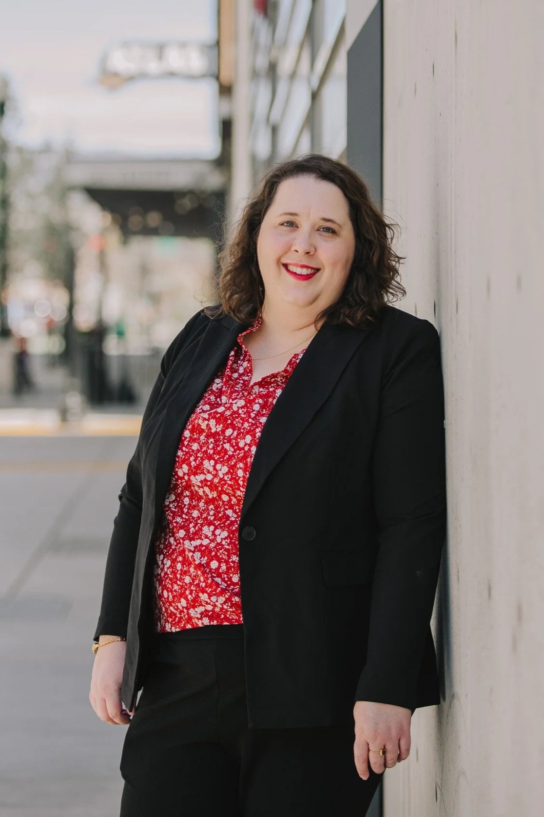 A woman with curly brown hair, wearing a black blazer over a red floral blouse, leaning against a white wall outdoors, smiling at the camera with a blurred city background.