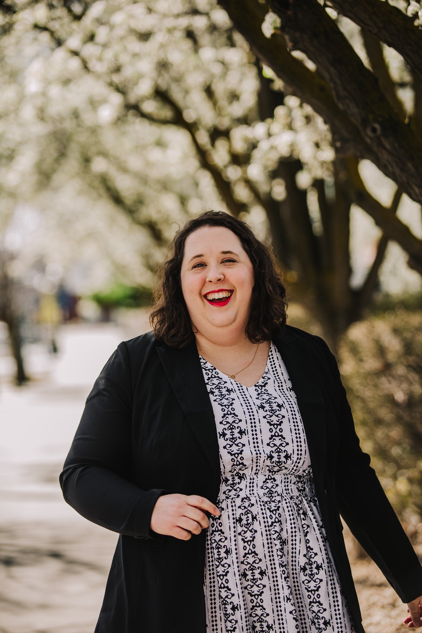A woman with dark curly hair smiling and laughing outdoors, wearing a black jacket over a white dress with black patterns, standing under a tree with white blossoms.