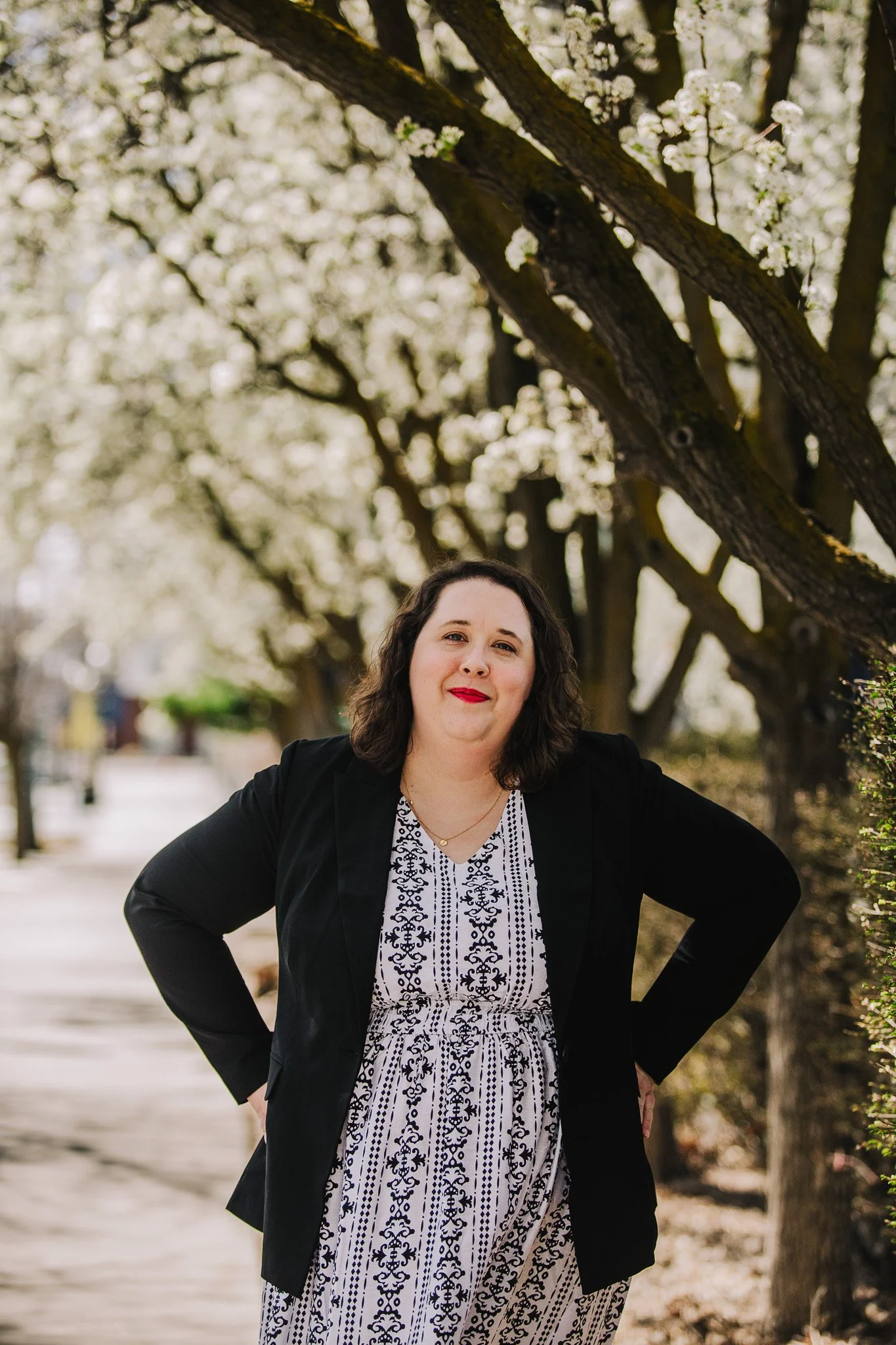 A woman standing outdoors on a pathway lined with trees blooming with white flowers, wearing a black blazer over a patterned dress.