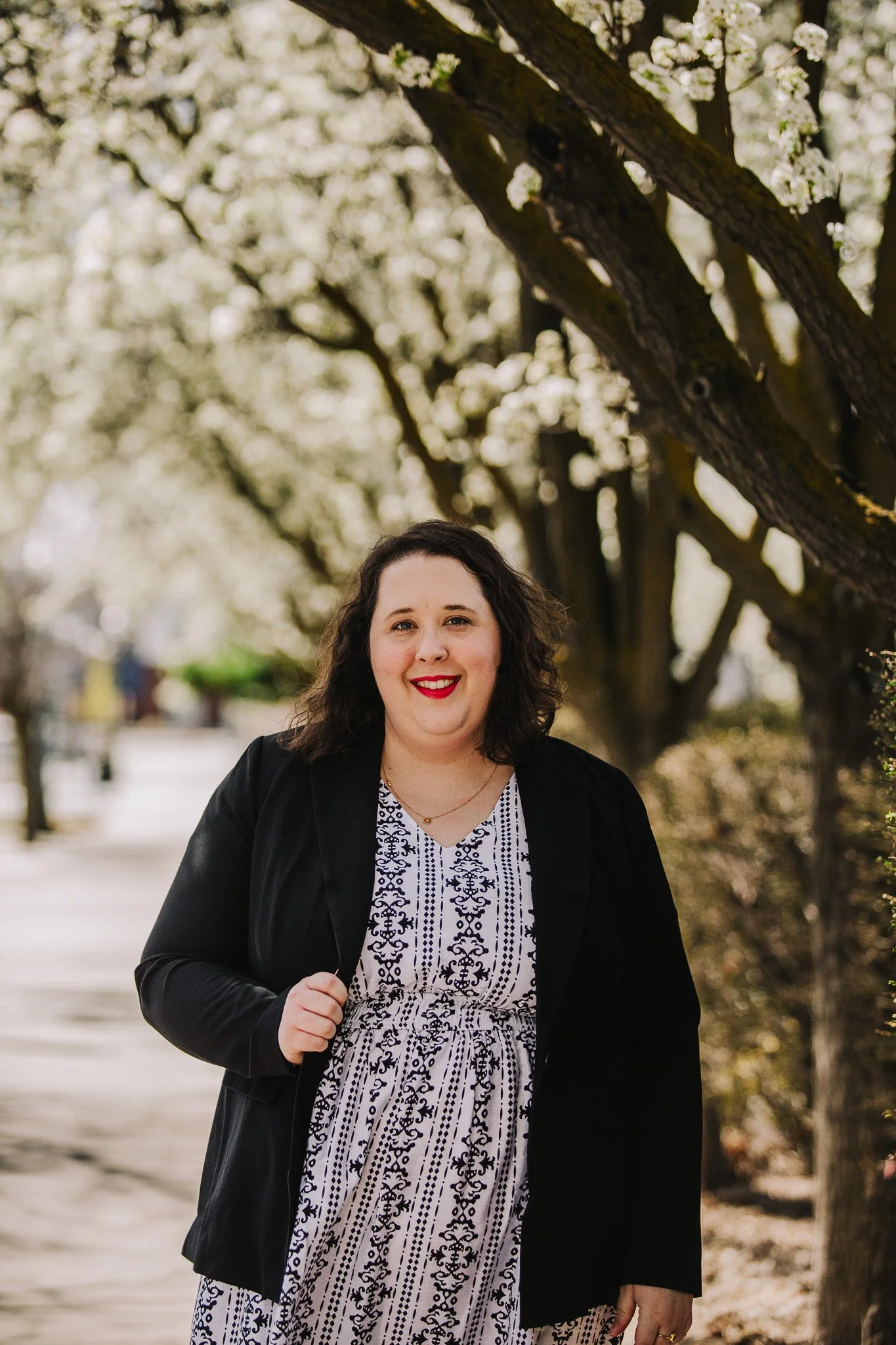 A woman with dark curly hair wearing a black jacket and a patterned dress, smiling outdoors during spring with blooming trees in the background.