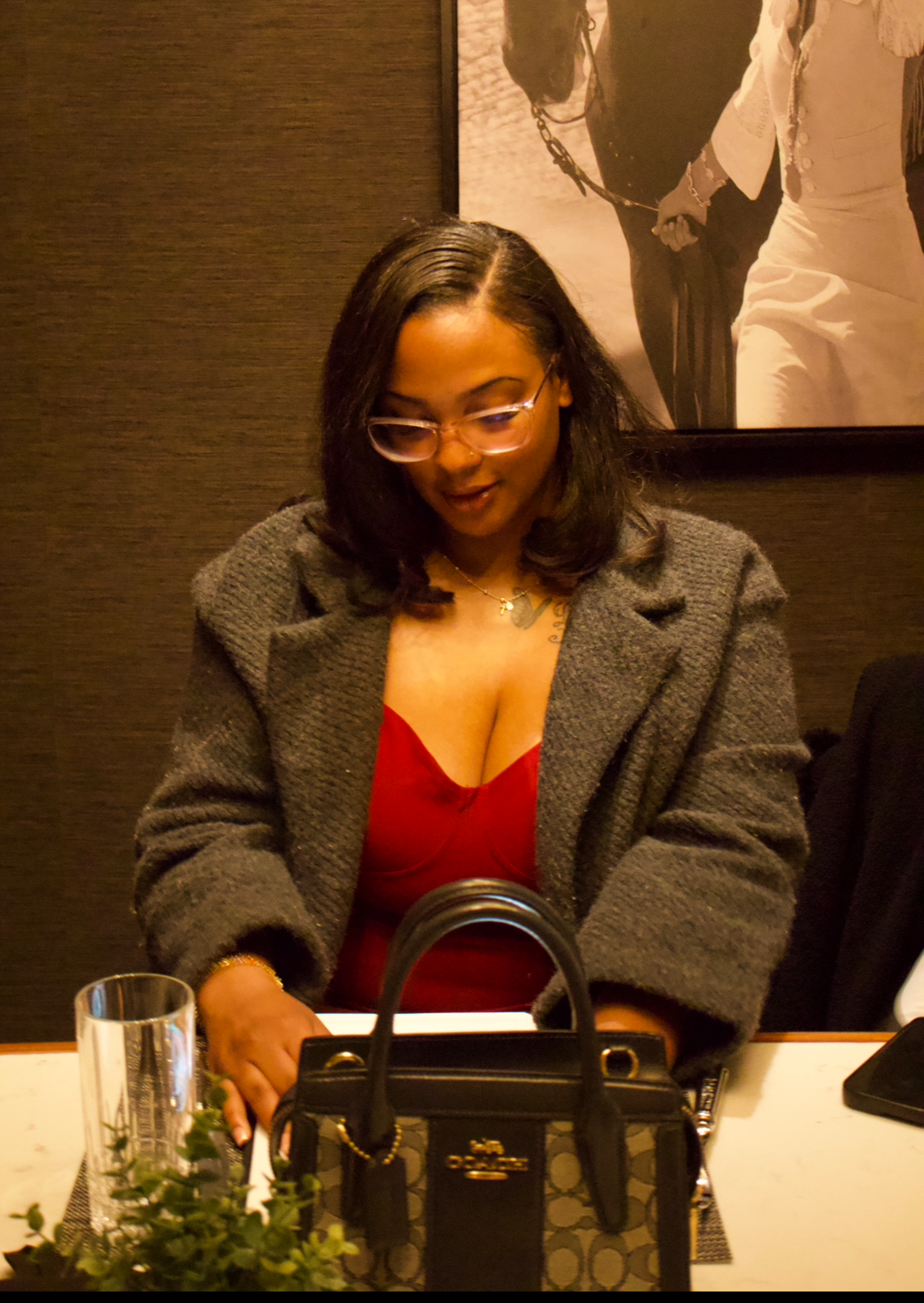 A woman with shoulder-length black hair and glasses, wearing a red top and a gray coat, sitting at a table with a black handbag, a glass, and some greenery, in a dimly lit restaurant or cafe.