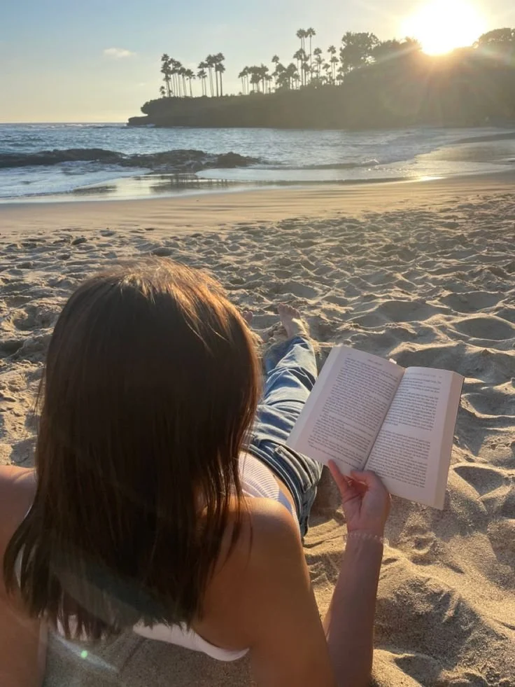 Woman lying on sandy beach, reading a book with ocean and palm trees in the background during sunset.