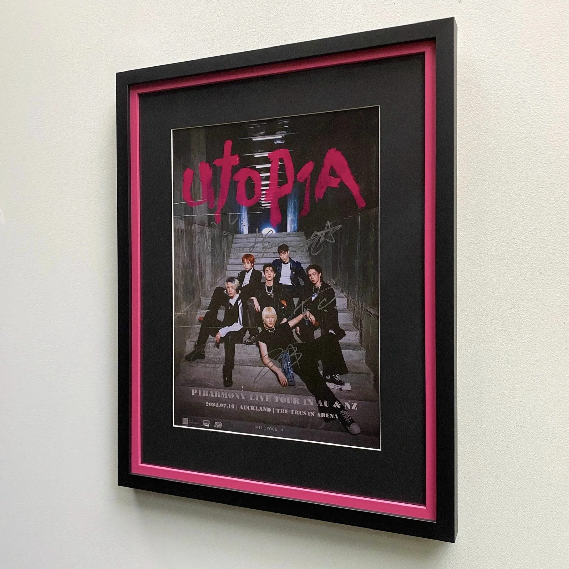 Framed poster of the band UTOPIA, featuring seven members in dark clothing sitting and standing on stairs against a gritty background, with pink handwritten style text and autographs, advertising a live tour at Auckland's Trusts Arena.