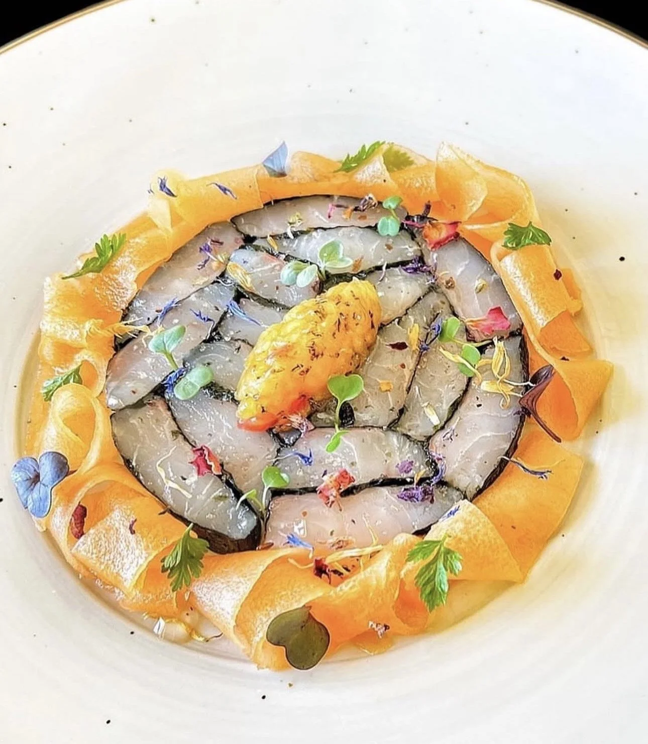 A plate of raw shrimp garnished with edible flowers, microgreens, and a central piece of orange-colored seafood or garnish, surrounded by a ring of orange peel ribbons.