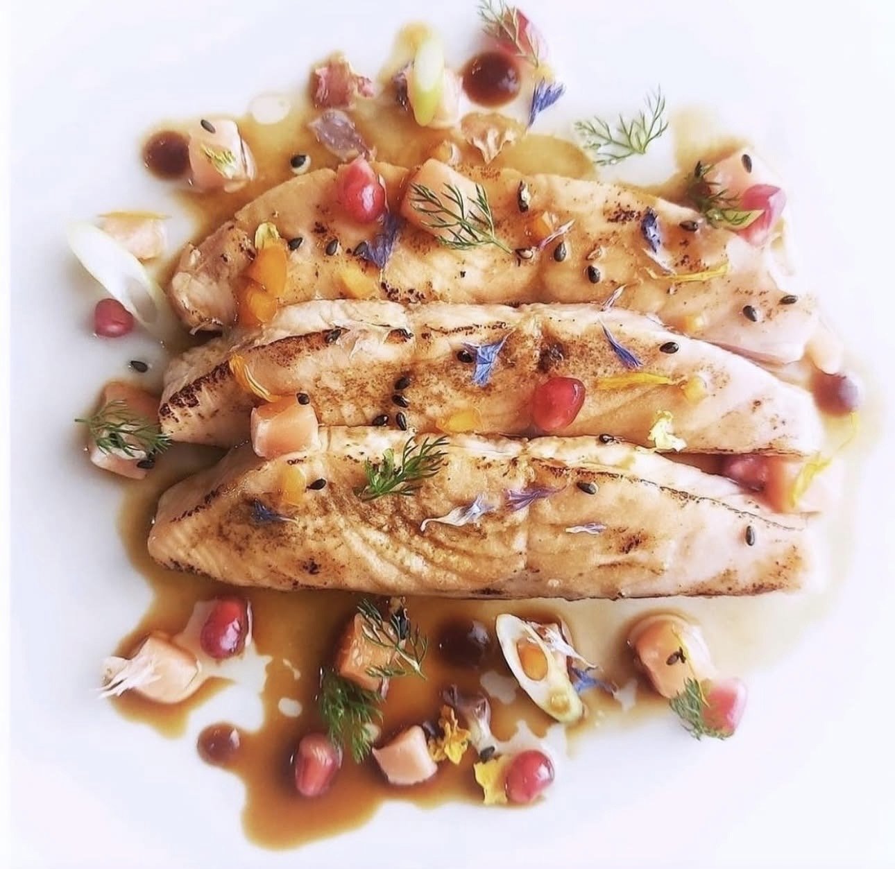Seared fish fillets garnished with herbs, pomegranate seeds, flower petals, and drizzled with sauce on a white plate.