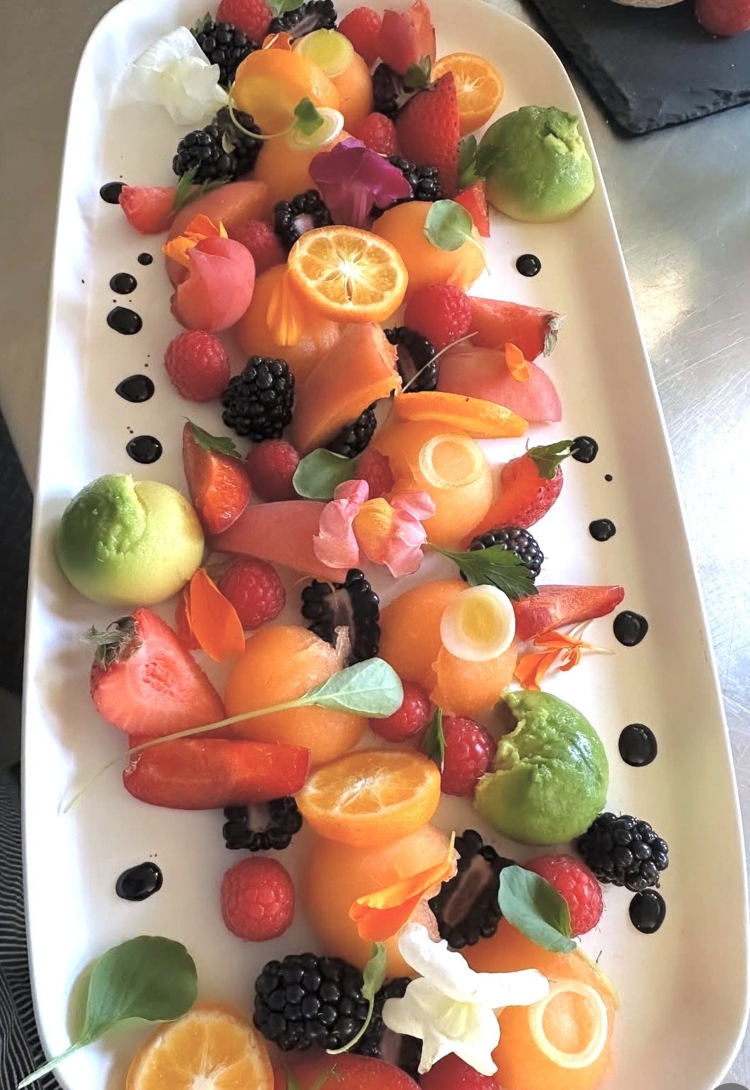 A white platter filled with a colorful variety of fresh fruits including strawberries, blackberries, raspberries, kumquat slices, a peeled green apple, and edible flowers, garnished with drops of dark syrup or balsamic reduction.