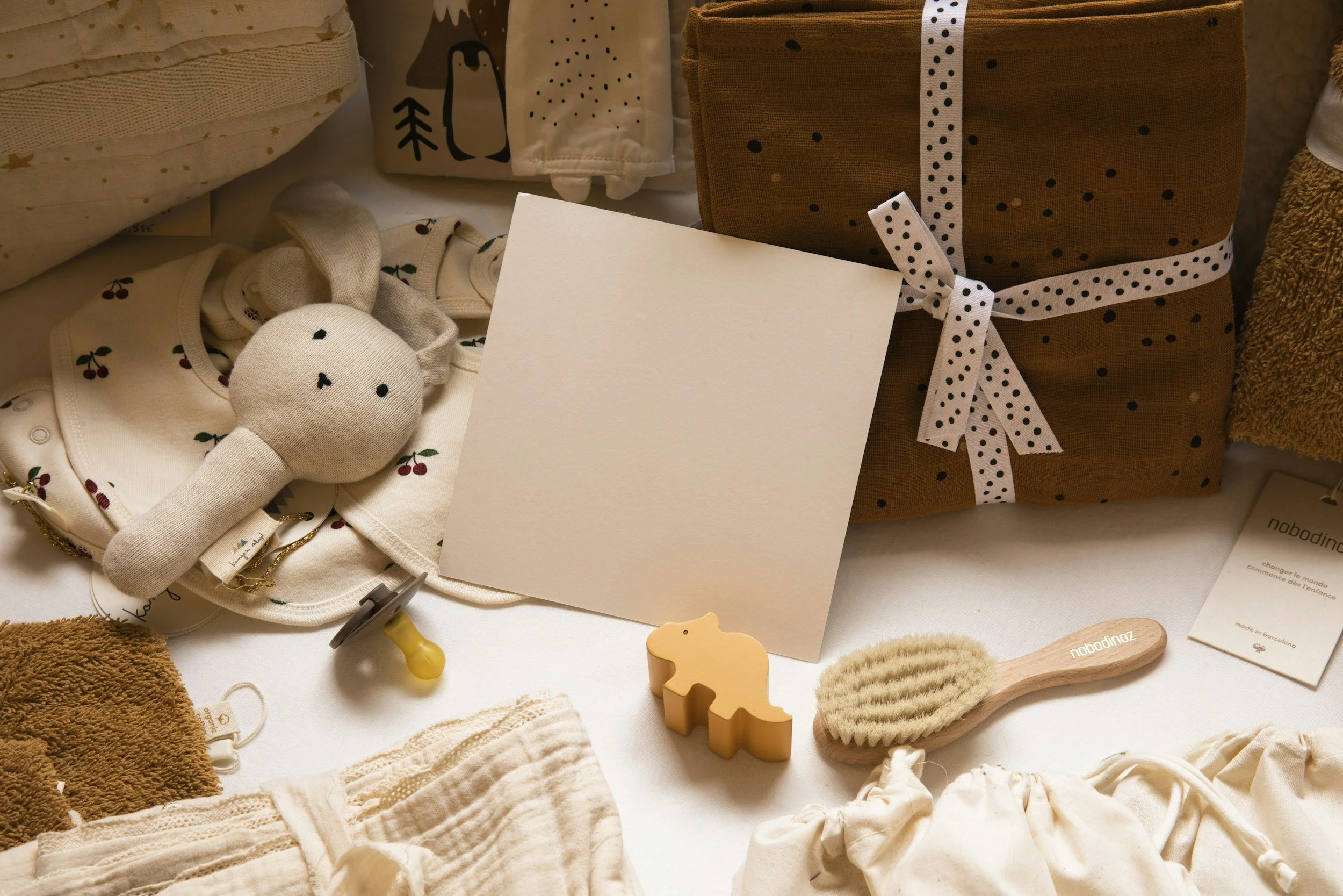 Baby clothes, a stuffed bunny toy, a gift box wrapped in brown paper with black polka dots and a white ribbon, a wooden rubber duck, a paddle brush, and various baby care items on a white surface.