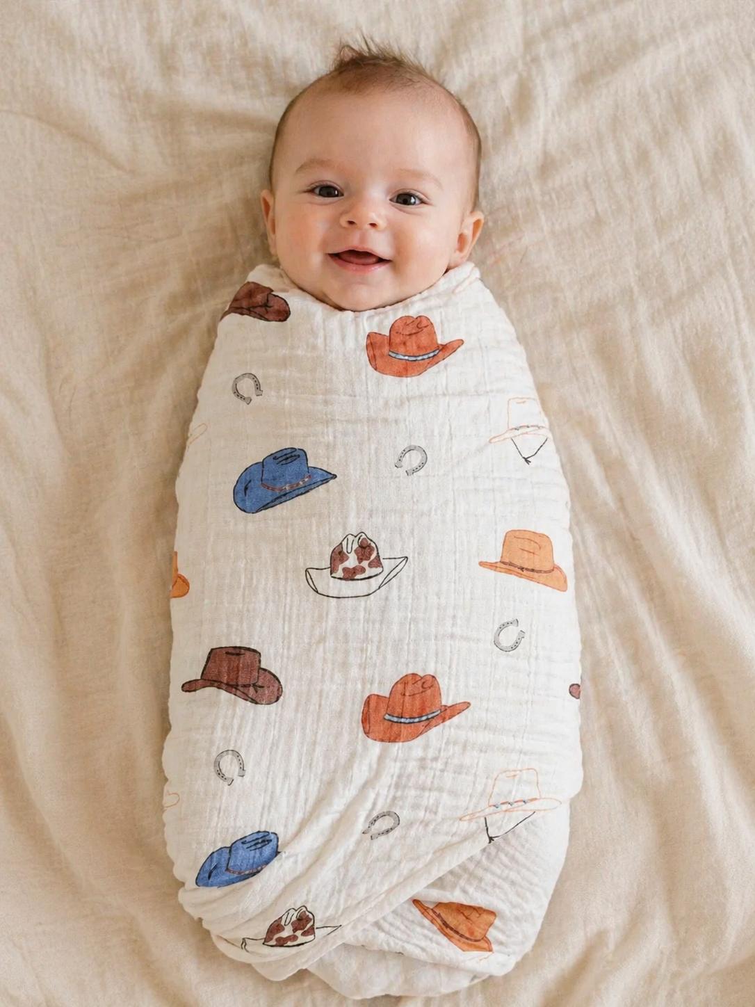 A smiling baby wrapped in a white swaddle blanket with cowboy hat and cake illustrations, lying on a beige blanket.