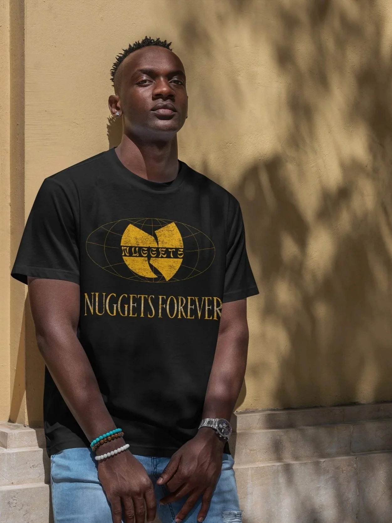 A young man with dark skin and short curly hair standing against a beige wall, wearing a black T-shirt with golden lettering and logo that reads "NUGGETS FOREVER" in the style of Wu-Tang Forever.