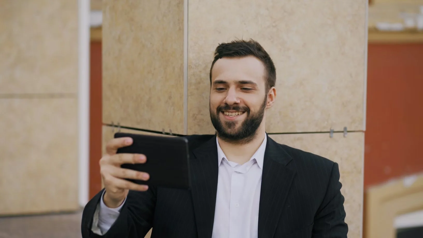 A man in a business suit smiling while taking a selfie with a smartphone in an indoor setting.
