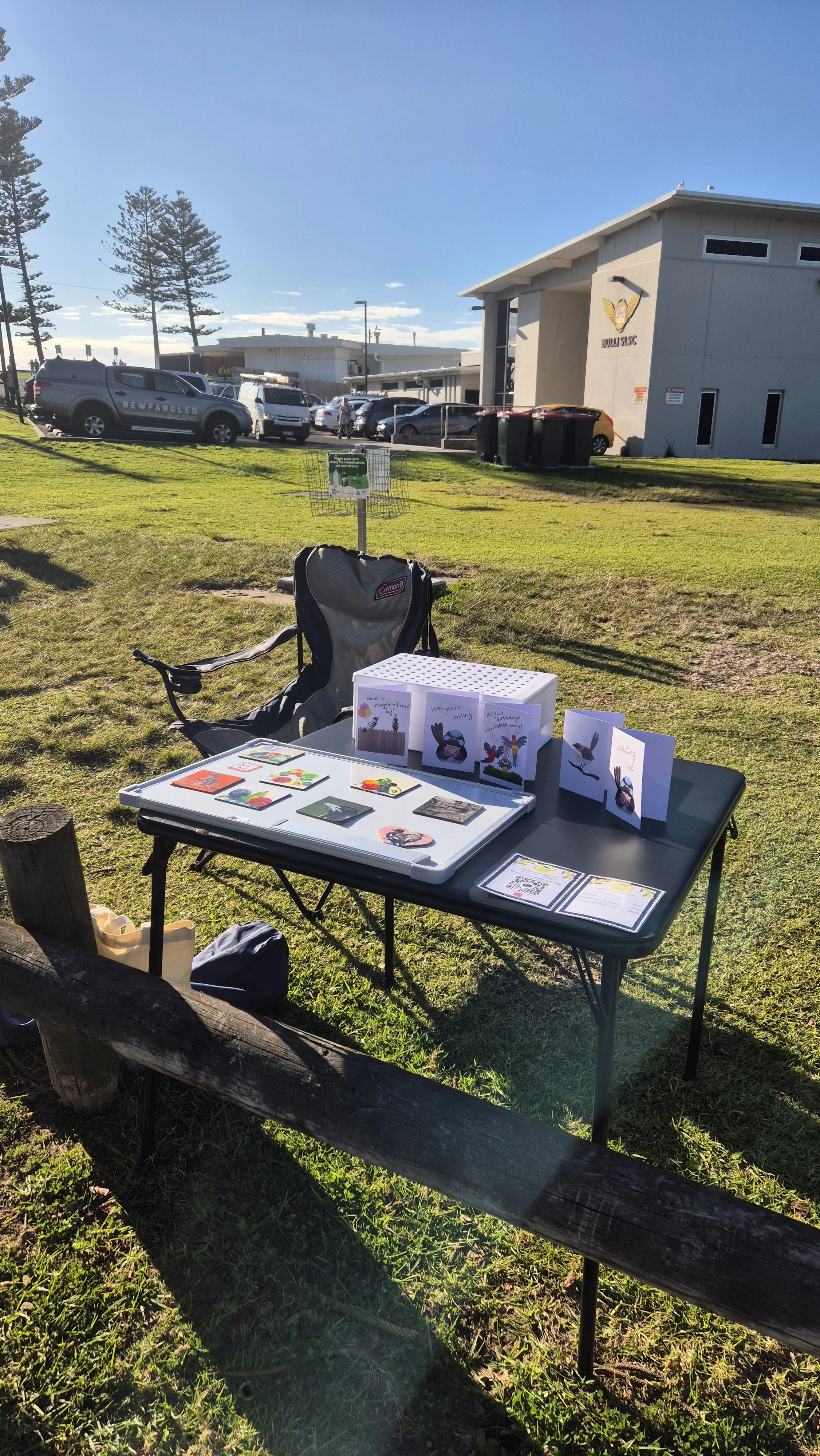 A fold out table set up on the grass with the Bulli Surf Life Saving Club building in the background. On the table is a tray of magnets, and 5 greeting card designs.