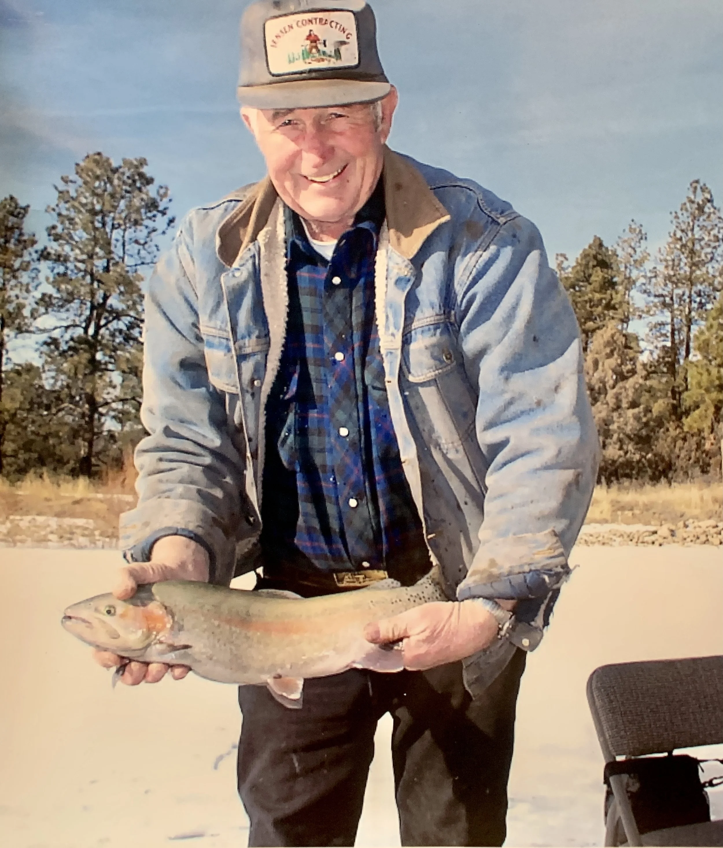 A man standing outdoors in a natural setting holding a fish with both hands. He is smiling and wearing a cap, a denim jacket, and a plaid shirt.