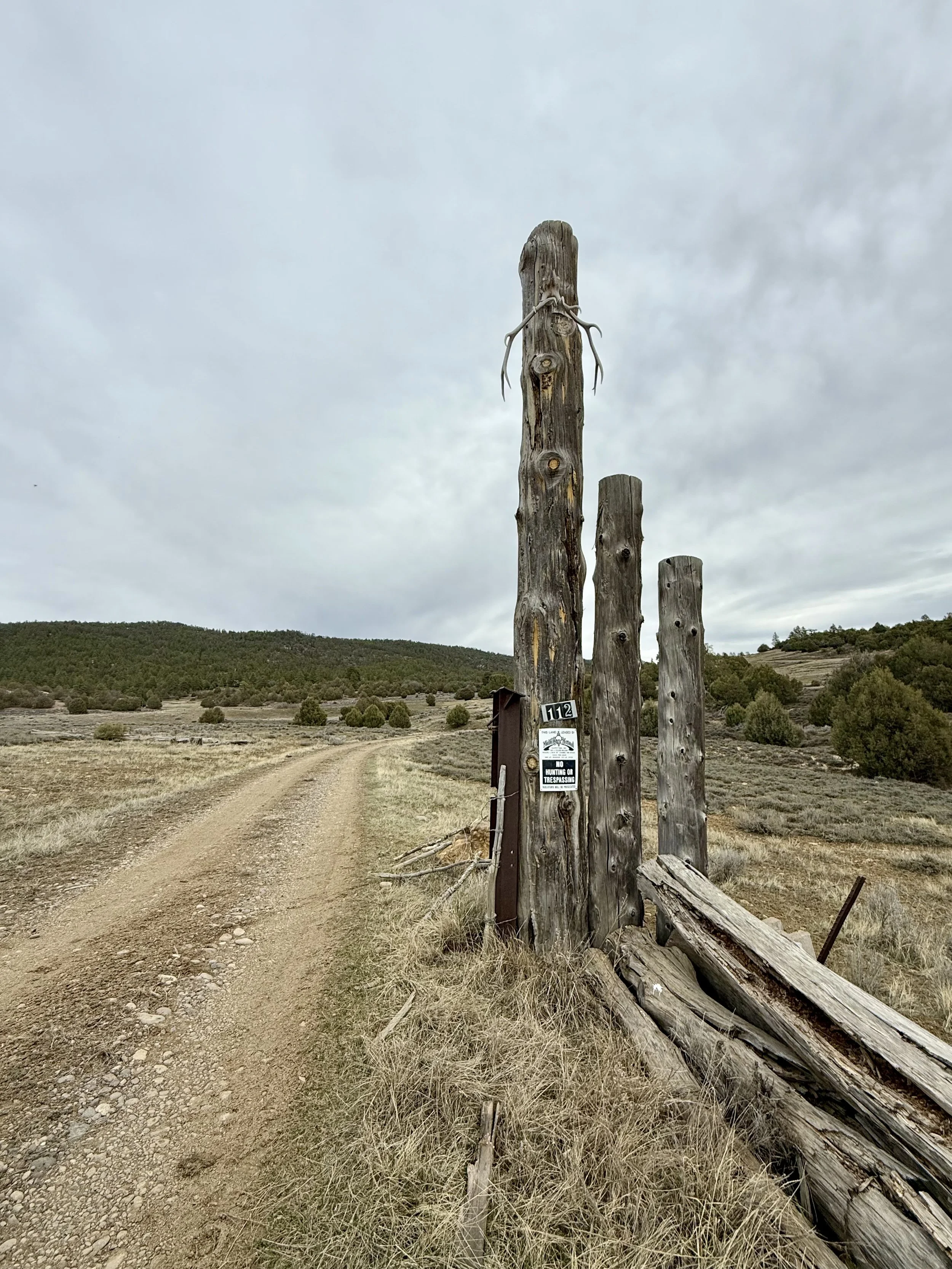 A dirt path through a rural landscape with tall wooden posts on the right, some leaning and weathered, with one post having a sign that reads 'No Hunting or Trespassing.' The background includes rolling hills and an overcast sky.