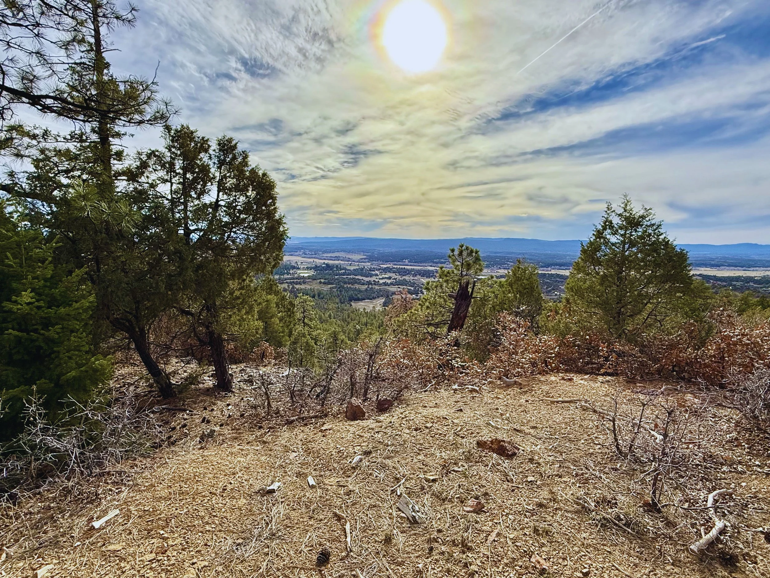 A scenic view from a mountain trail overlooking a vast valley with scattered buildings, trees, and distant mountains under a partly cloudy sky with the sun shining brightly.