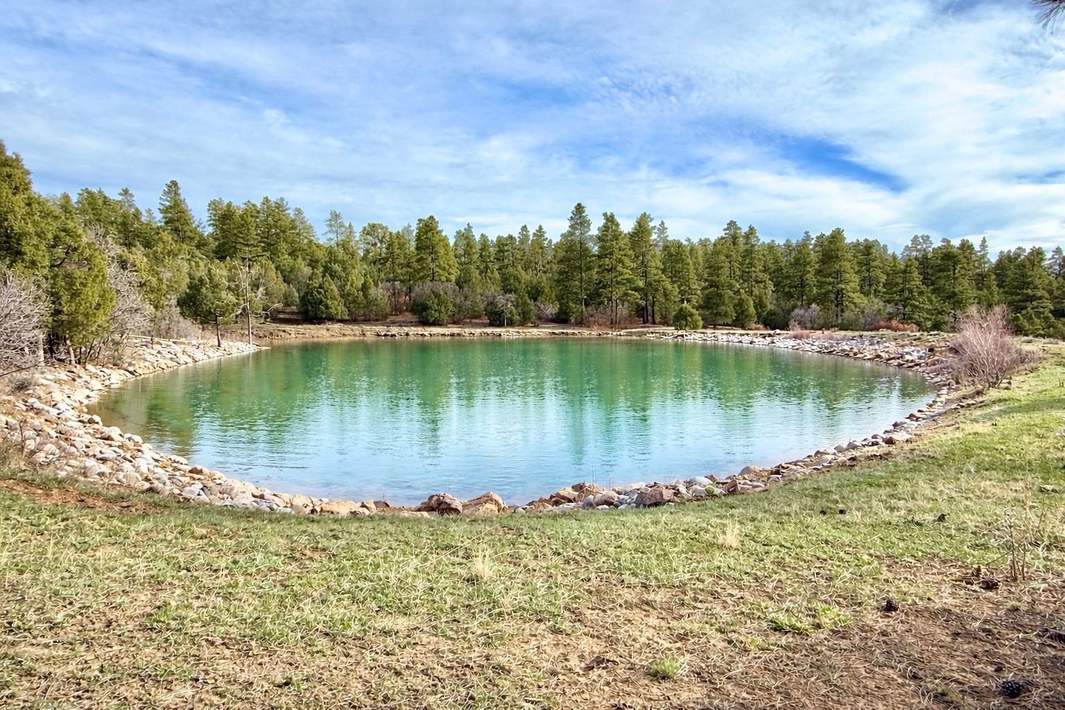 A small, clear green lake surrounded by rocks and green pine trees under a partly cloudy blue sky.