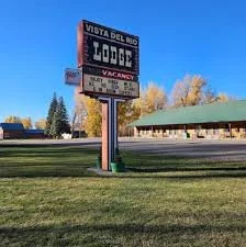 A roadside sign for Vista del Rio Lodge and Motel with vacancy indication, on a grassy area with trees and a building in the background under a clear blue sky.