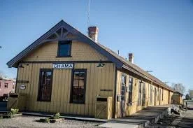 A yellow wooden train station building with a sign that reads 'Ghana'; it has a gabled roof, several windows, and is located on train tracks with a clear blue sky in the background.