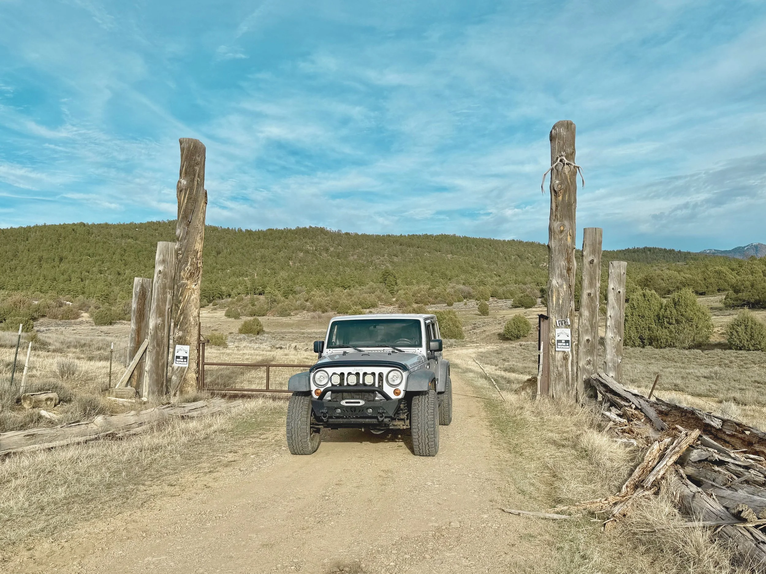 A Jeep parked in front of a rustic wooden gate made of weathered logs, with a hilly landscape and green trees in the background under a partly cloudy sky.