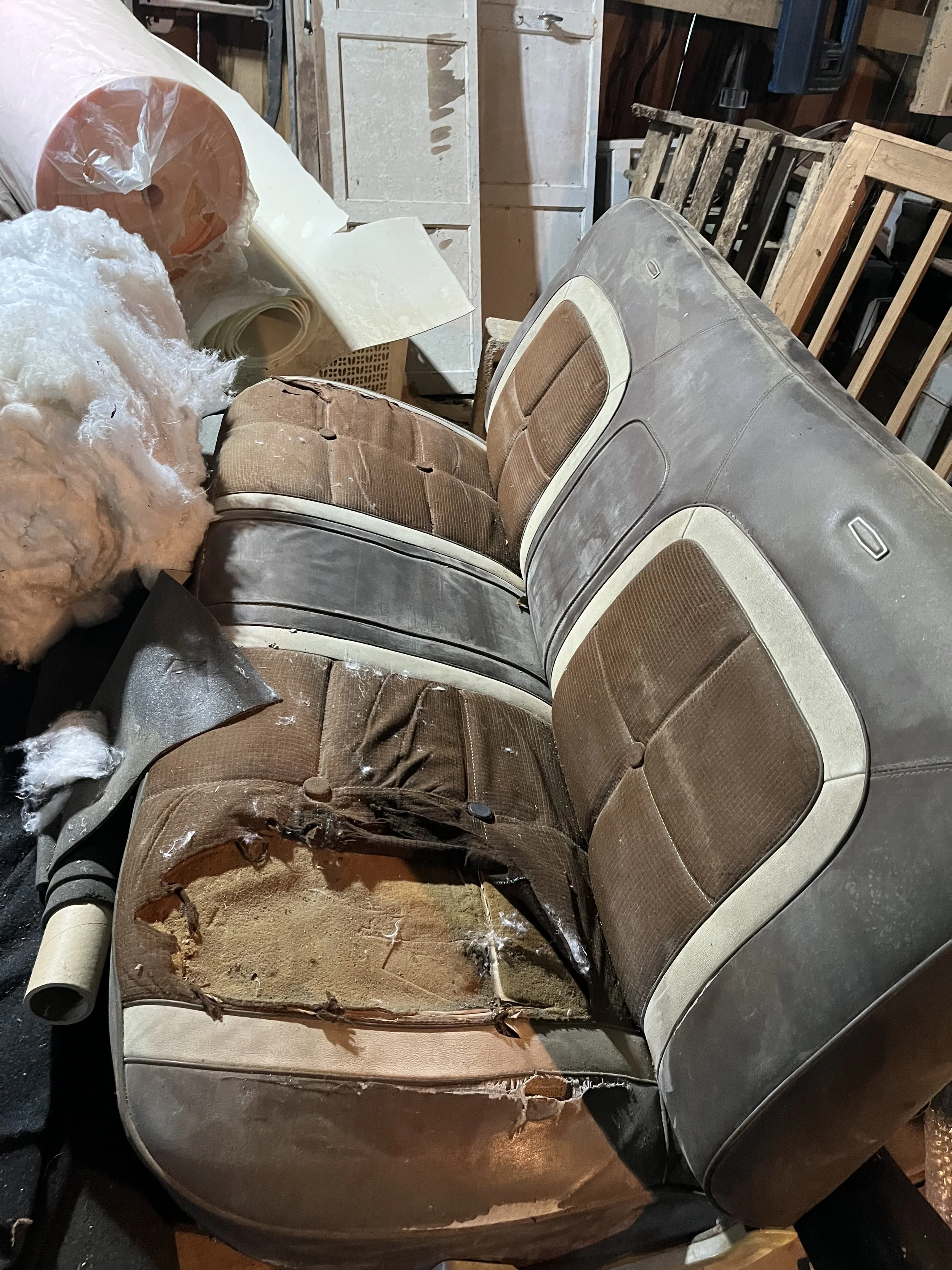 Old, damaged car bench seat with torn upholstery and exposed foam, surrounded by various debris in a cluttered garage.