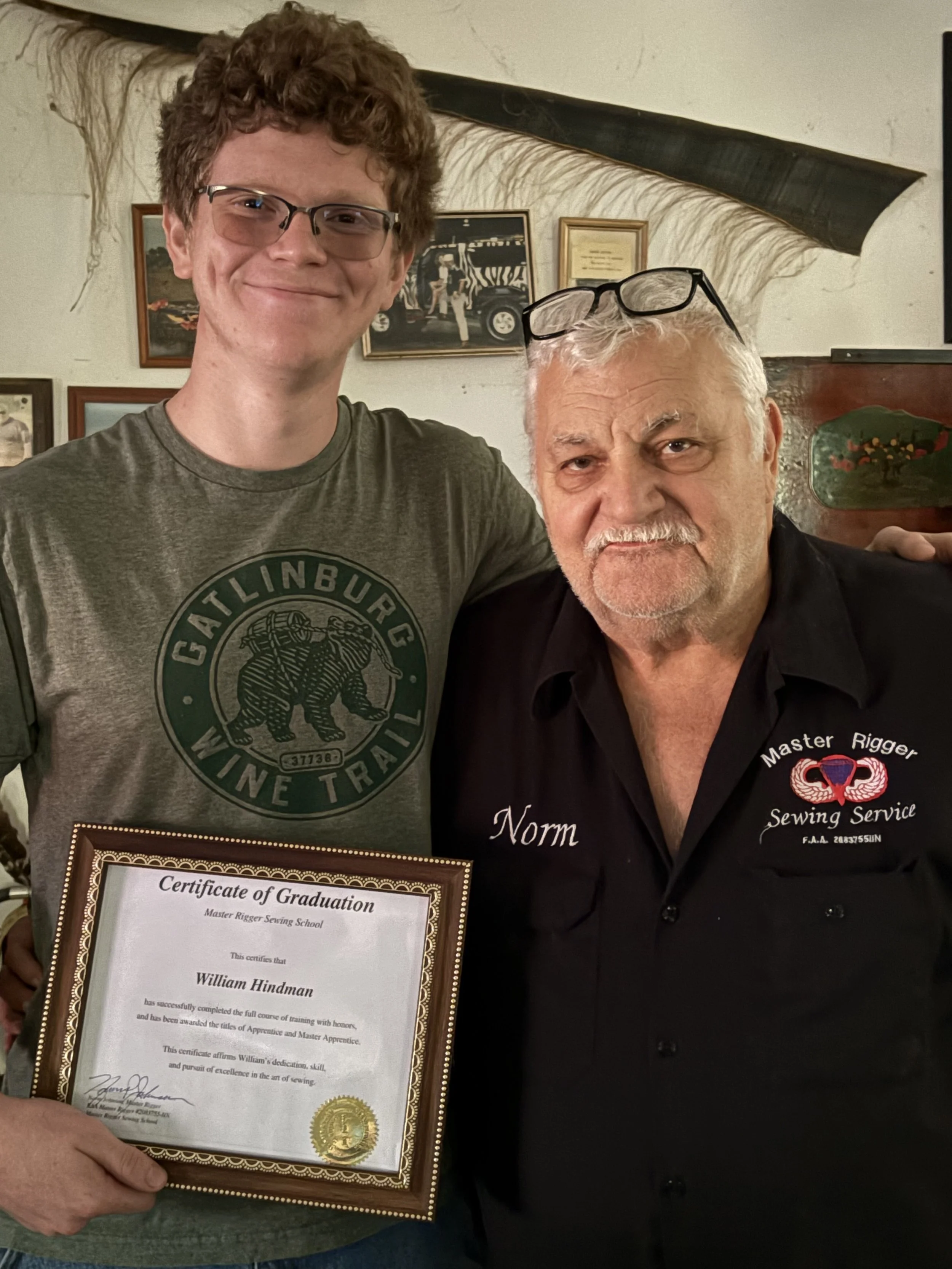 A young man and an older man are posing together, with the young man holding a certificate of graduation. The younger man is wearing glasses and a gray t-shirt with a graphic of a bear and the words 'Calinburg Wine Trail'. The older man, possibly his instructor or mentor, is wearing a black shirt with embroidery that reads 'Master Rigger Sewing Service' and 'Norm', and has glasses resting on his forehead. They are smiling and standing in a cozy room decorated with framed pictures and paintings.