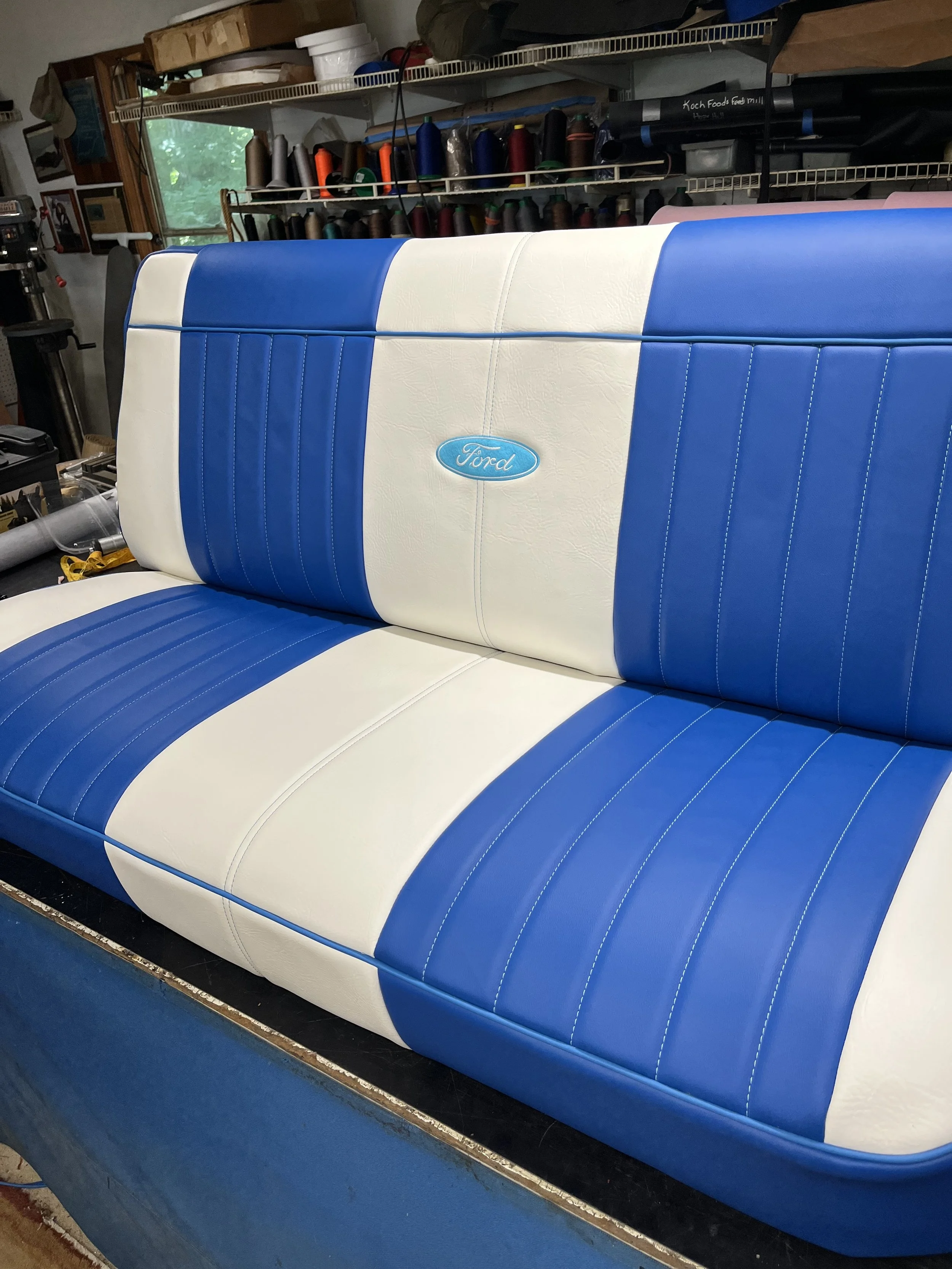 A vintage-style car bench seat with blue and white leather upholstery, featuring the Ford logo embroidered on the middle section.