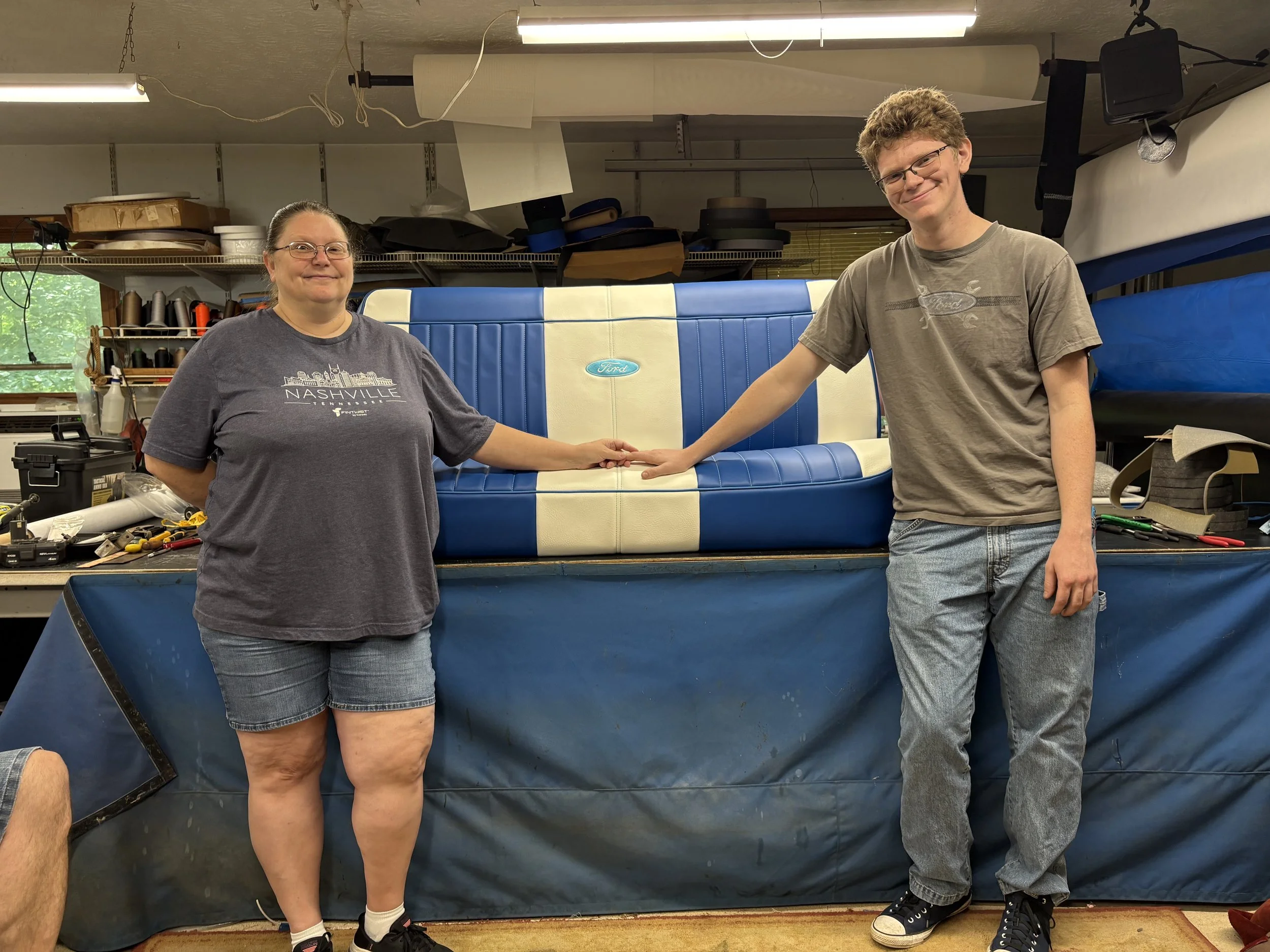 A woman and a young man in a workshop, holding hands over a blue and white Ford logo on a vintage car upholstery. The woman wears glasses and a gray T-shirt with 'Nashville' printed on it, and shorts. The young man wears glasses and a gray T-shirt with a Ford logo, jeans, and sneakers. The workshop has tools, shelves, and supplies in the background.