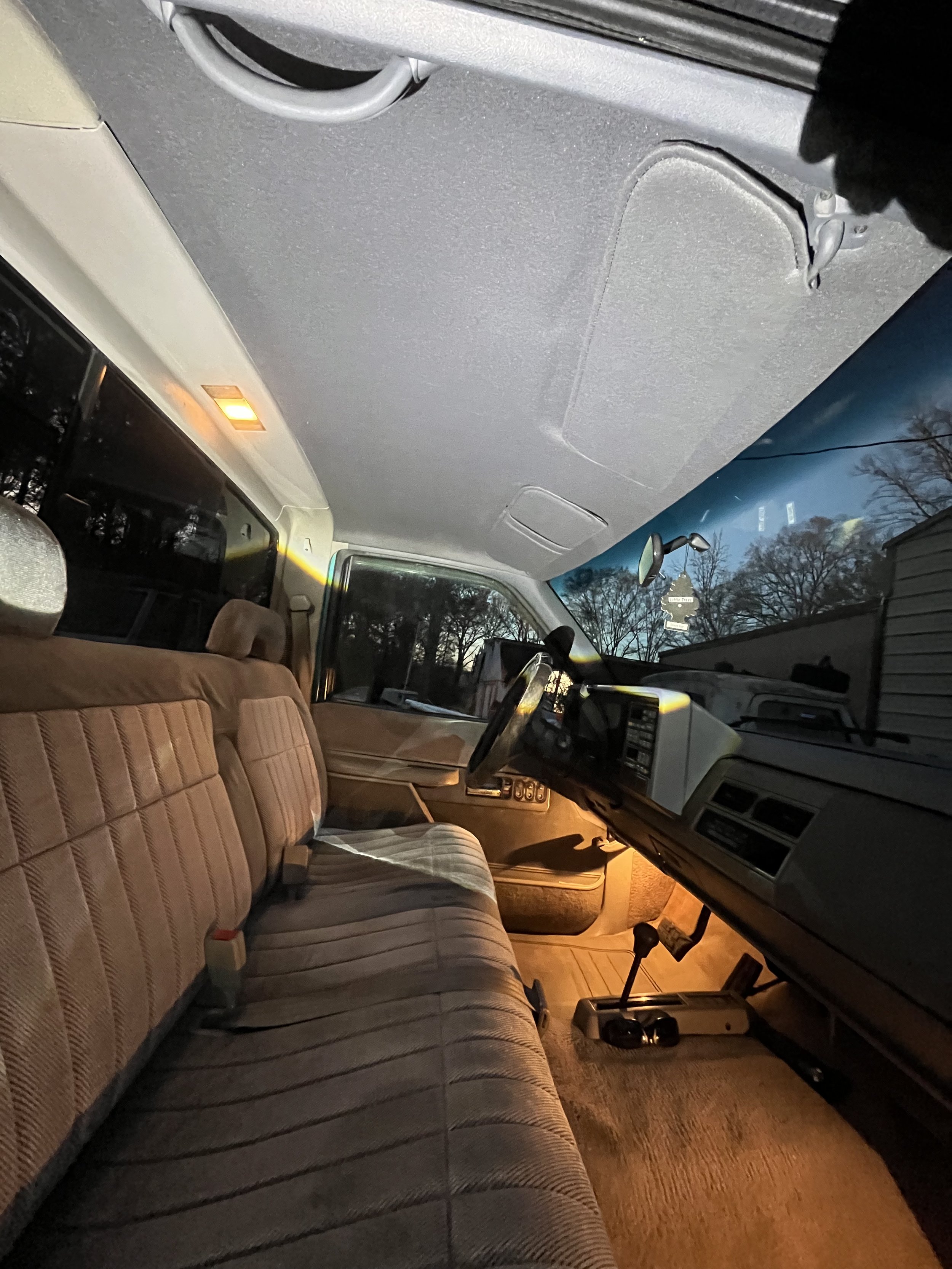 Interior of an old vehicle with a tan fabric bench seat, dashboard, steering wheel, gear shift, and overhead light, taken during dusk or early evening.