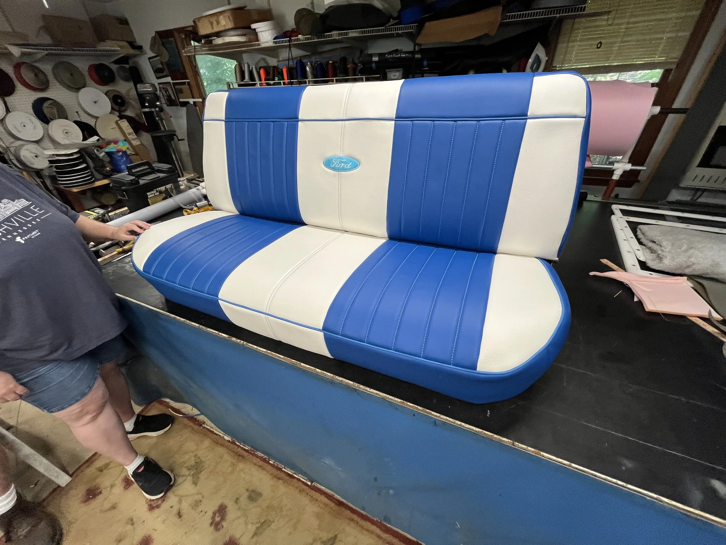 A vintage Ford car bench seat with blue and white upholstery inside a workshop or garage.