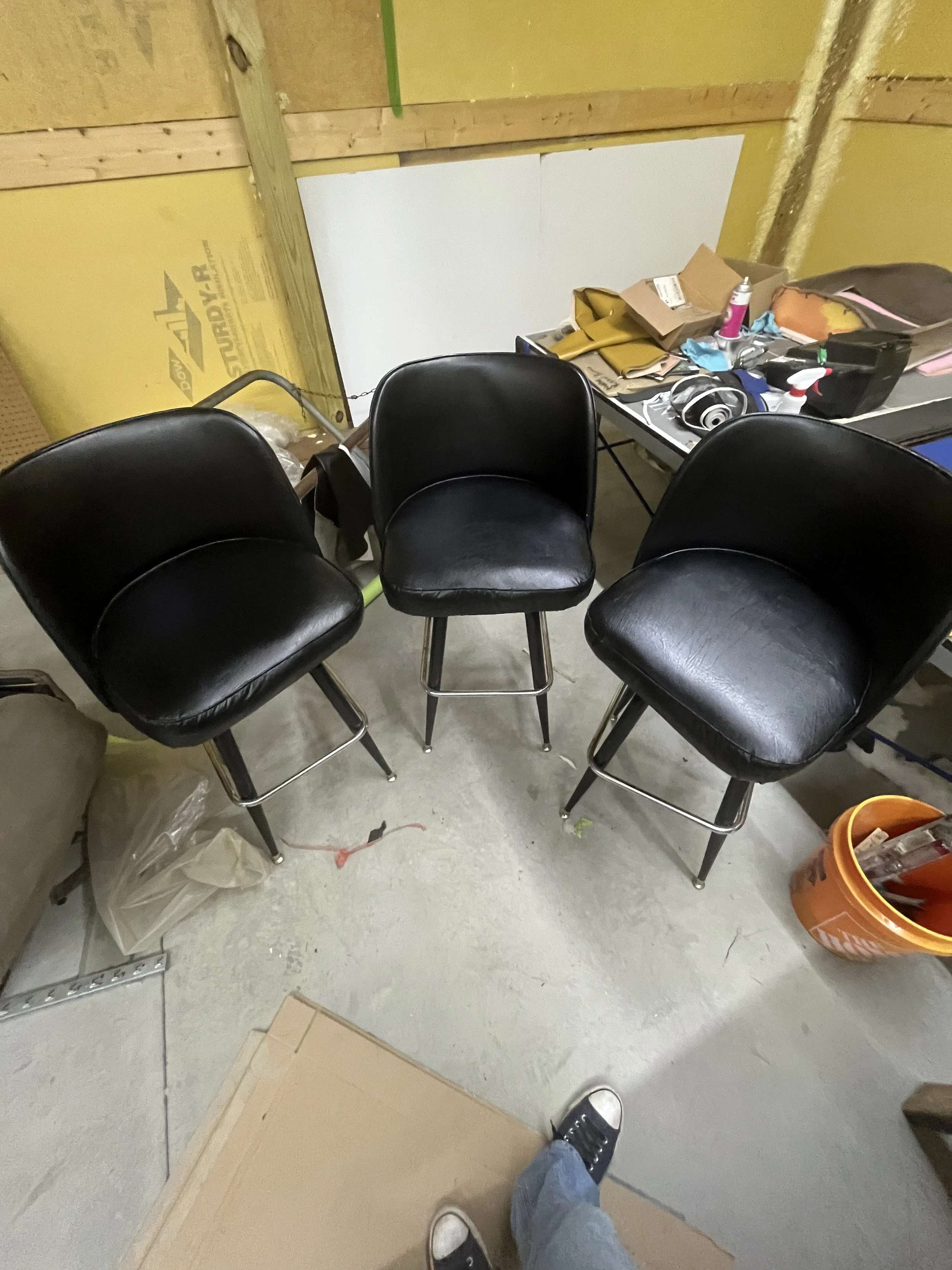 Three black bar stools with chrome legs are arranged in a semi-circle facing the camera in a workshop or garage setting. In the background, there is a cluttered table with paper, bottles, tools, and headphones, and yellow insulation and wooden framing on the wall.