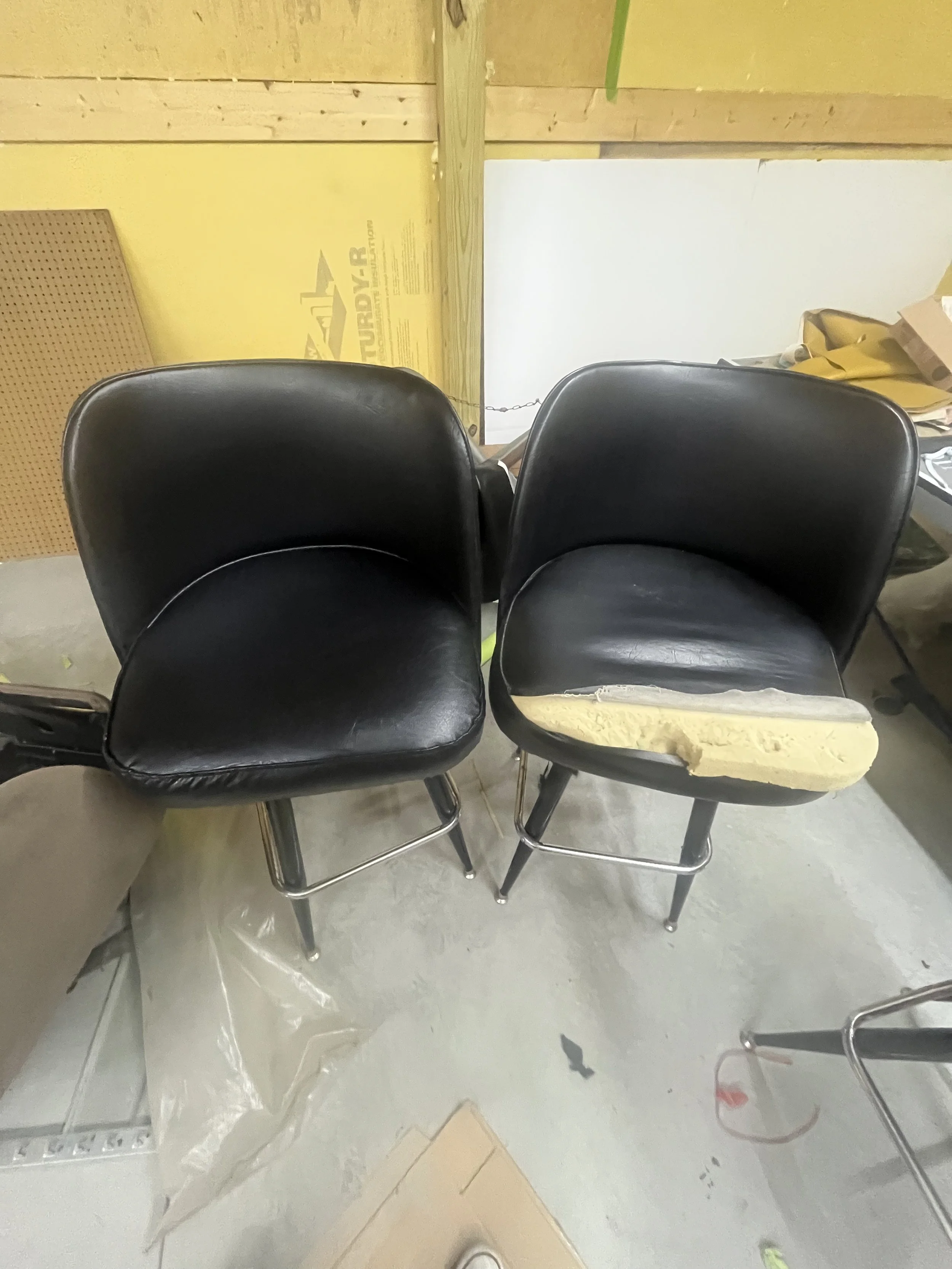 Two black vintage-style chairs with metal legs, one chair has a damaged seat cushion with foam padding exposed. They are placed in a workshop or storage area with plywood walls, cardboard, and yellow insulation material.