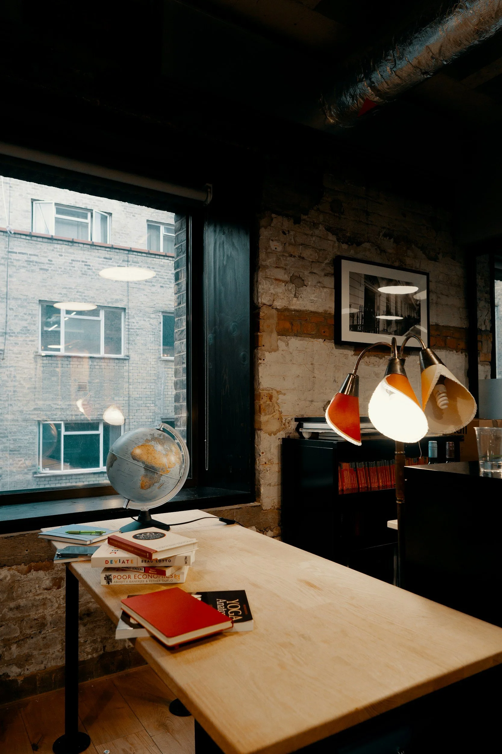 A cozy office space with a wooden desk, a globe, a stack of books, and a lamp with red, yellow, and white shades. The room has exposed brick walls and large windows with a view of an adjacent building.