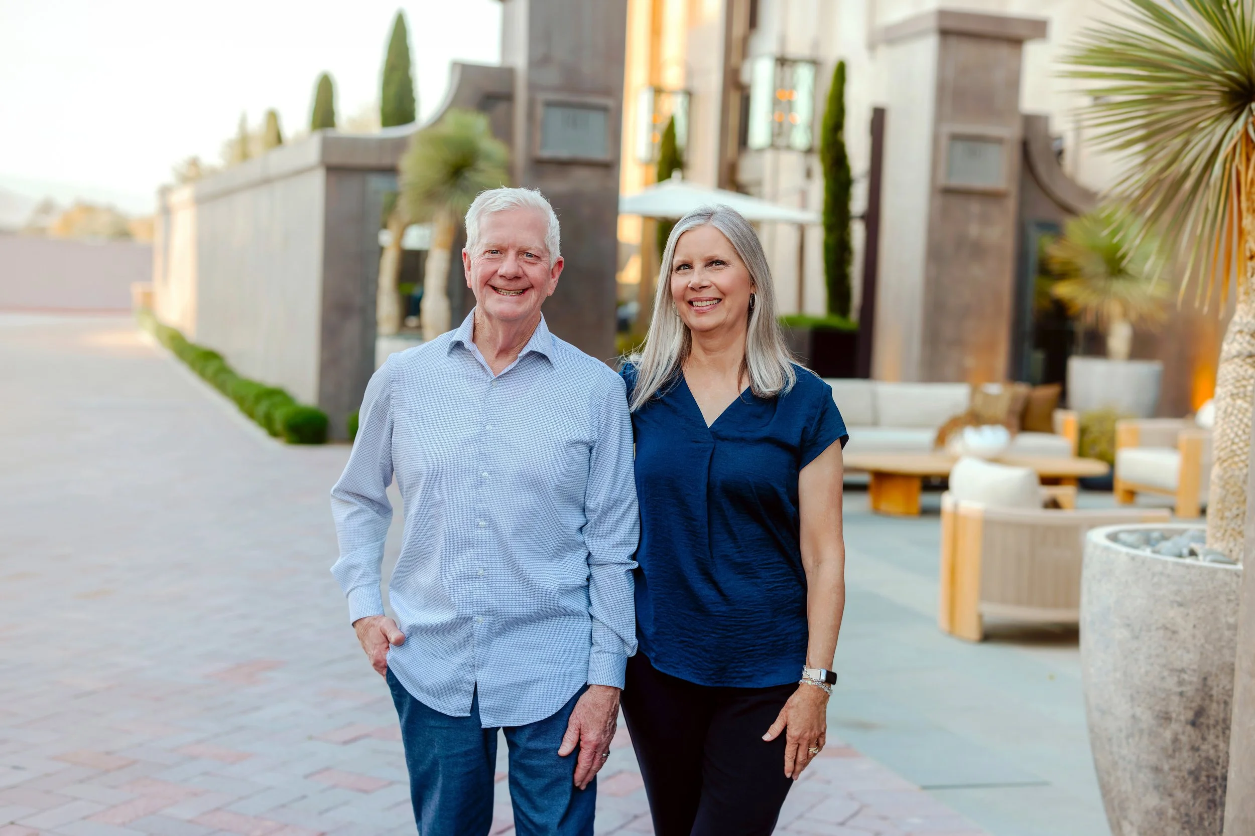An elderly couple standing outside in a well-maintained patio area with modern outdoor furniture and potted plants, smiling at the camera.