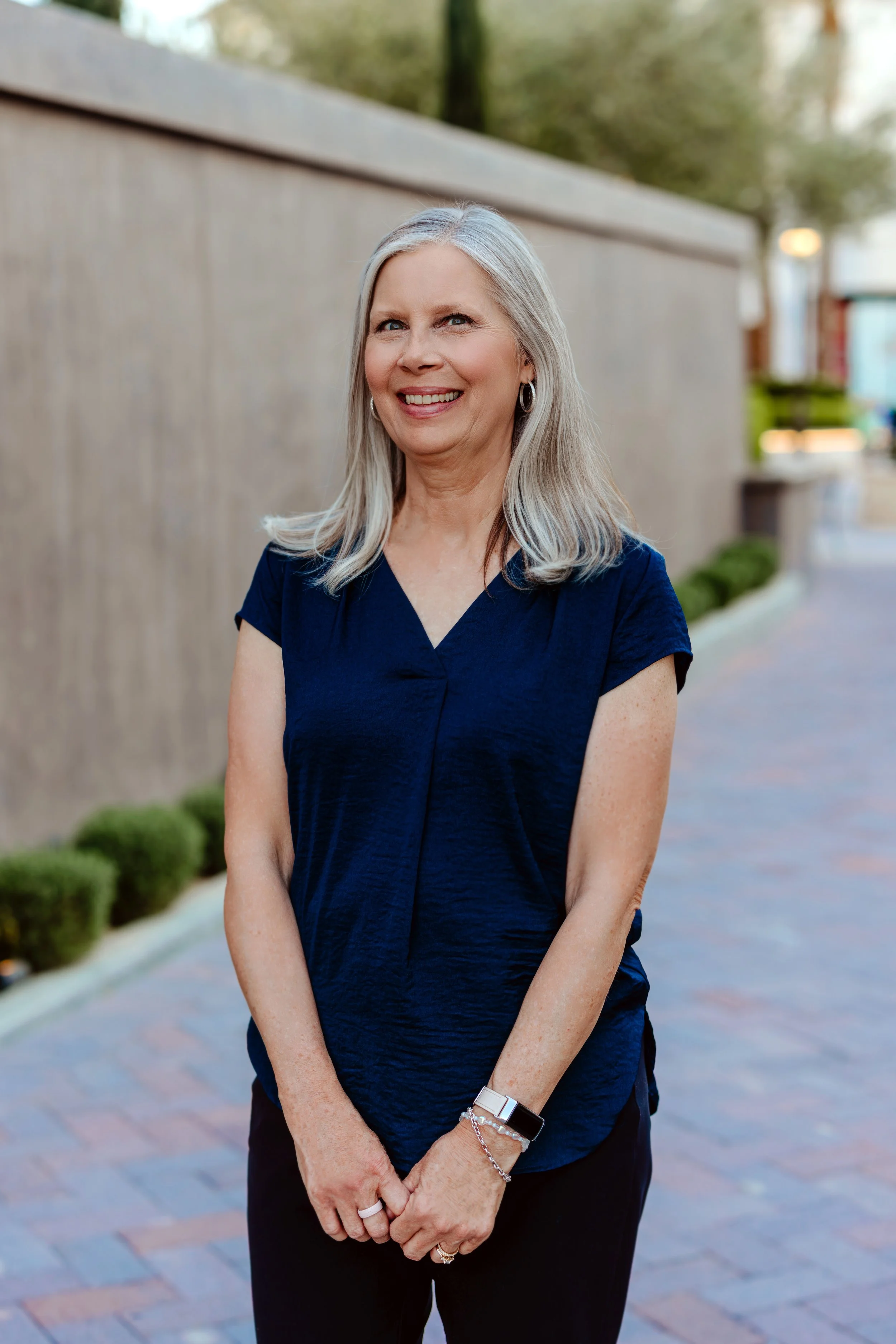 A middle-aged woman with long gray hair, wearing a navy blue blouse and black pants, standing outdoors on a brick sidewalk with a beige wall and greenery in the background, smiling at the camera.
