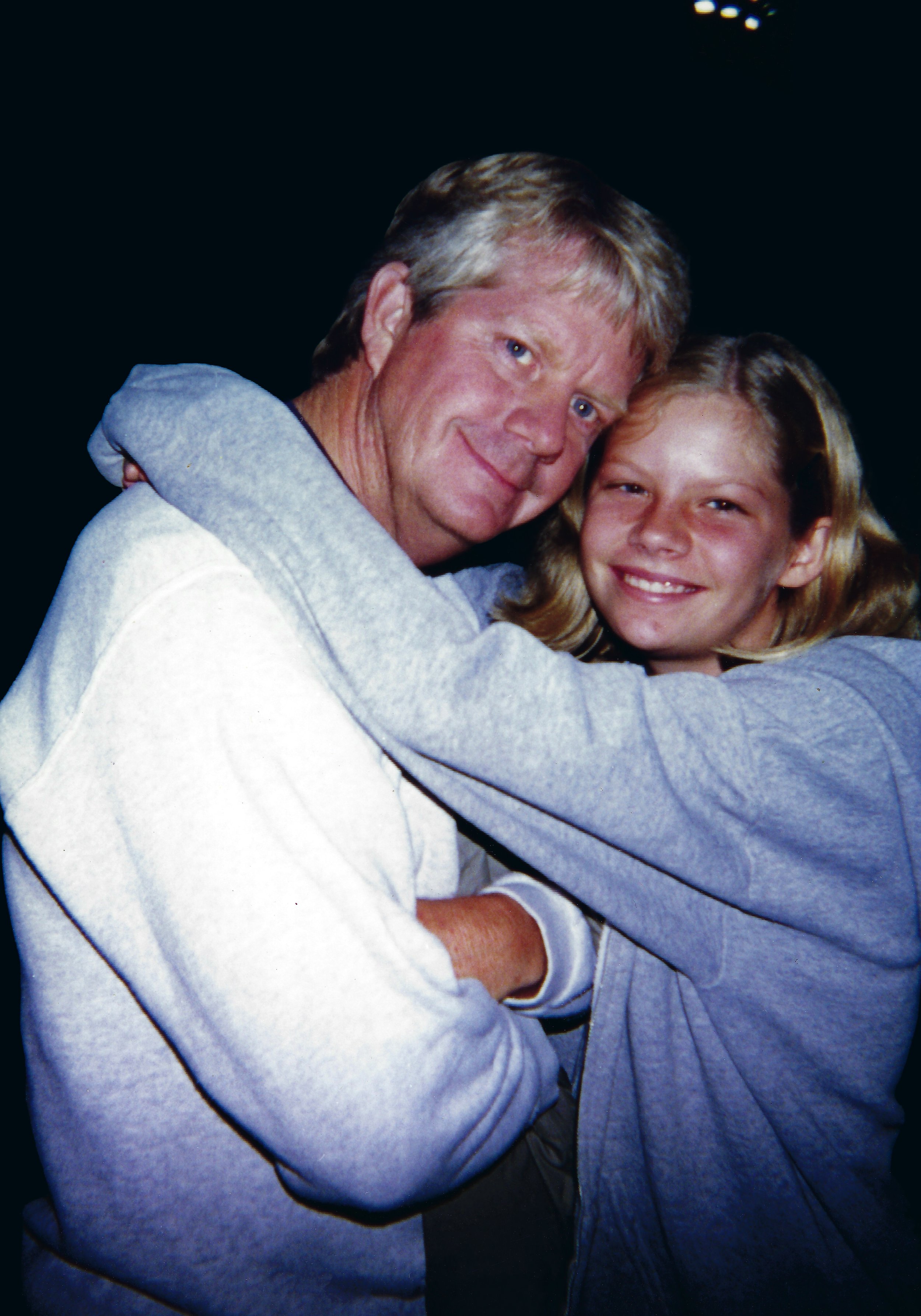 A man and a young girl embracing each other, both smiling at the camera, with the man wearing a white sweatshirt and the girl wearing a gray hoodie.