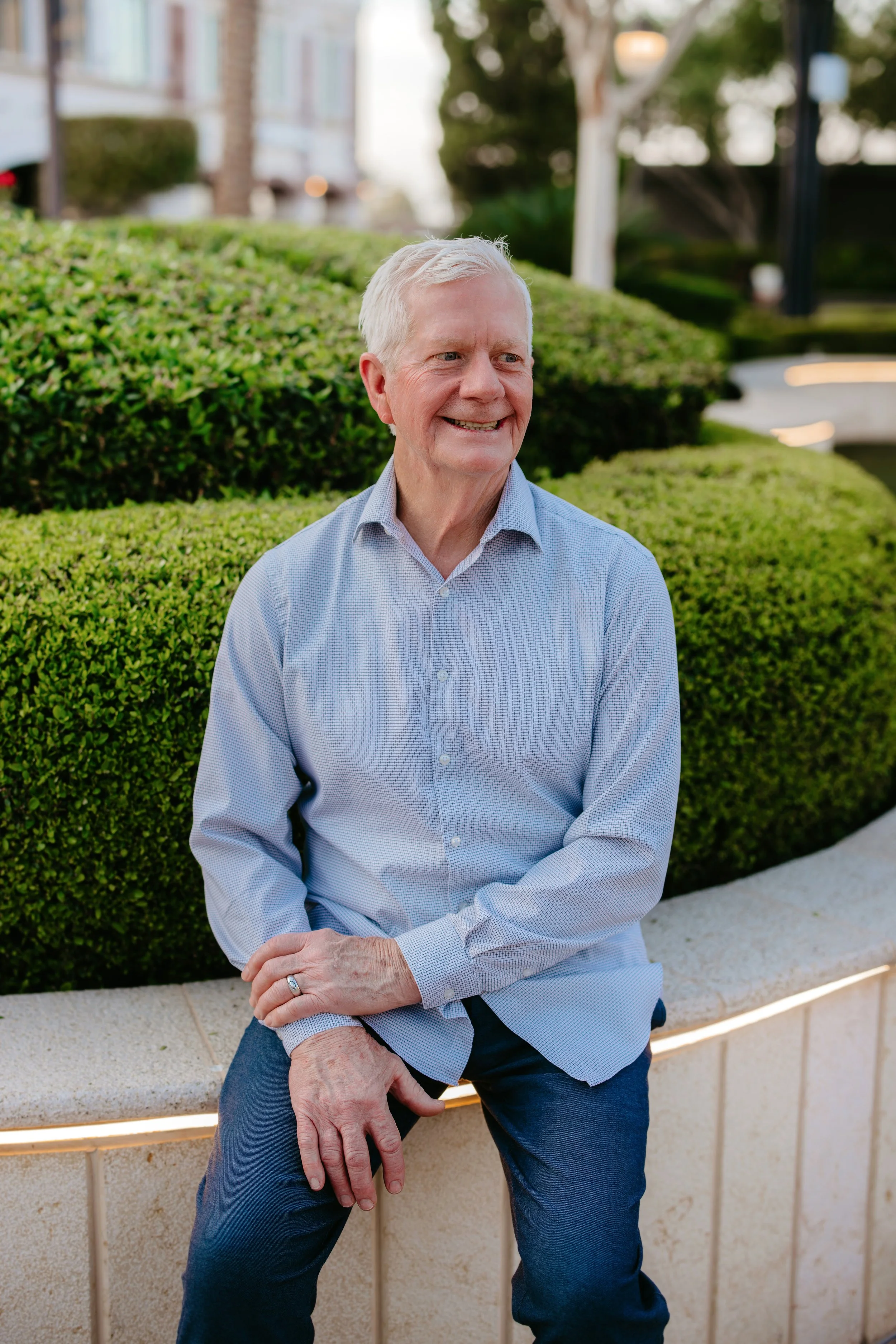 An elderly man with white hair sitting on a curved concrete bench in a park, smiling. He is wearing a light blue button-up shirt and dark blue pants, with a ring on his left hand. There are well-maintained bushes, trees, and buildings in the background.