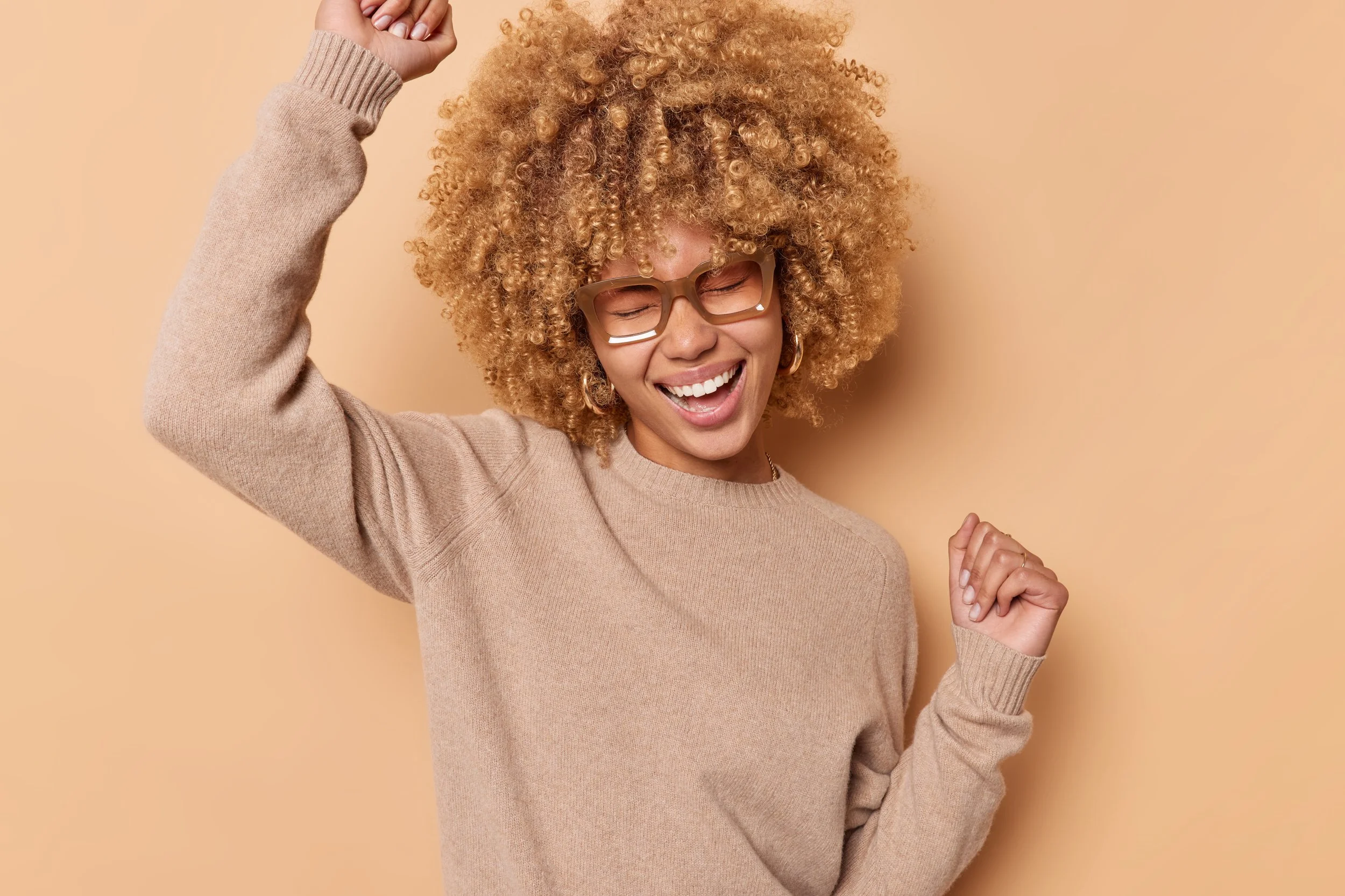 A woman with curly blonde hair, wearing glasses, a beige sweater, smiling and dancing with her eyes closed against a light peach background.