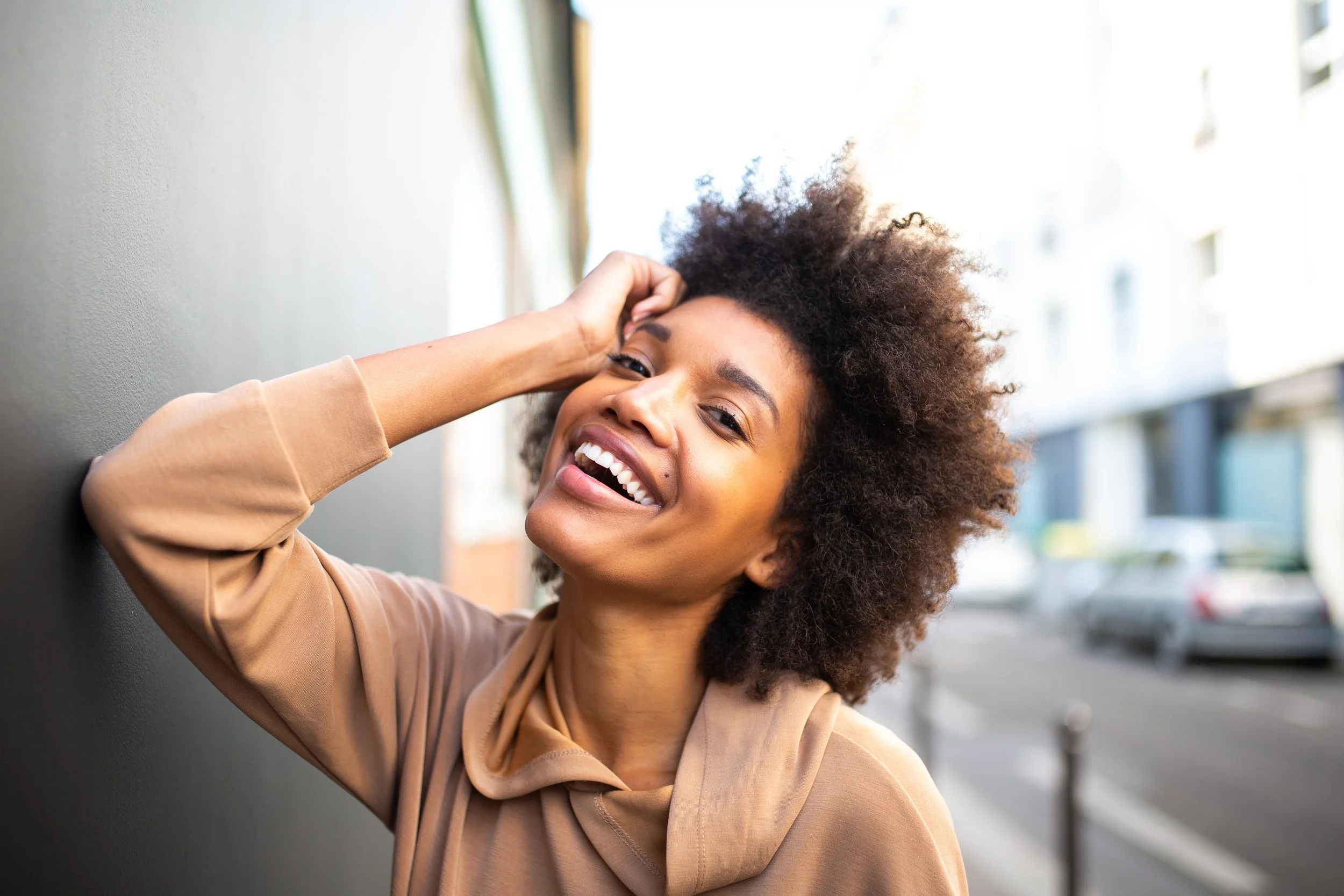 A woman with curly hair leaning against a wall, smiling and holding her head with one hand.