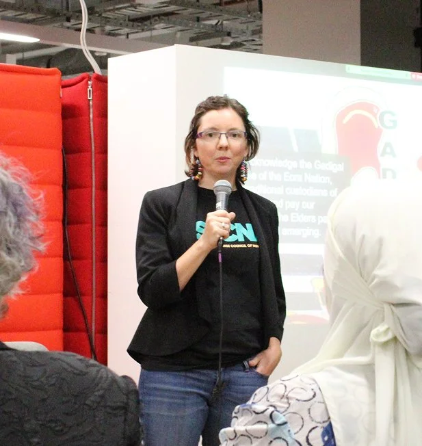 Kylie (CEO of SECNA) in a SECNA t-shirt and blazer speaks into a microphone at an event, with a slide acknowledging the Gadigal people of the Eora Nation visible on screen behind her.