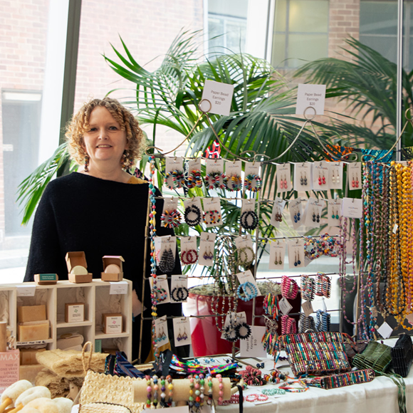 A woman stands behind a colourful market stall displaying handmade jewellery including beaded earrings, bracelets and necklaces, alongside wooden gift products.