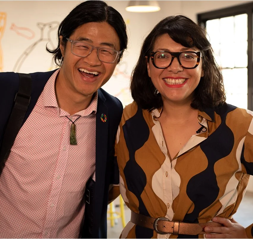 Two SECNA members smiling and laughing together at an indoor event — a man in a pink shirt and navy blazer, and a woman with dark hair and bold-framed glasses.