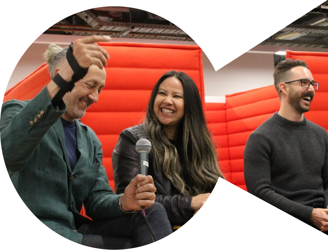 Three panellists laughing together at a SECNA event, seated in front of a red backdrop — a woman holds a microphone while the man beside her raises his hand animatedly.