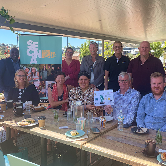 A group of ten people gathered around an outdoor table at a SECNA community meeting, with one person holding up a document showing partner logos, and a SECNA banner visible in the background.