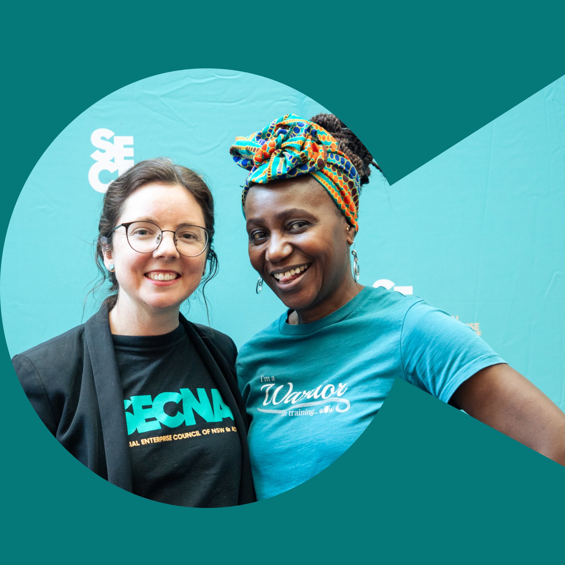 Two women smiling at the camera, in front of a media wall with the SECNA logo. The woman on the left wears a SECNA t-shirt, the woman on the right wears a shirt that reads "I'm a Warrior in training"