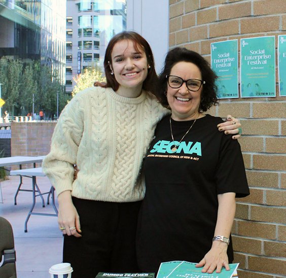 Two women smiling together outdoors at a Social Enterprise Festival, one wearing a SECNA t-shirt, with a brick wall and event signage in the background.