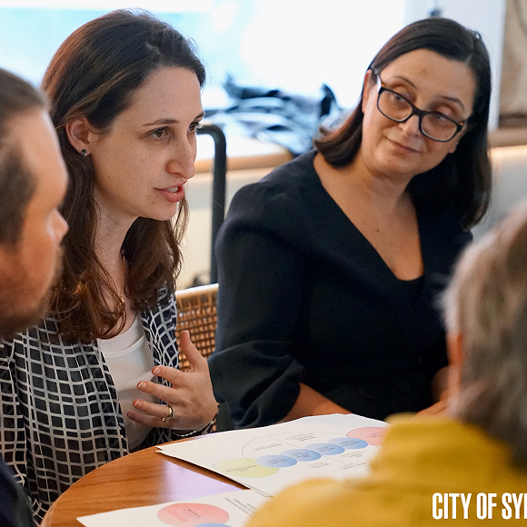 A woman speaks animatedly to a small group around a table, with a diagram document visible in the foreground, while another woman listens attentively.
